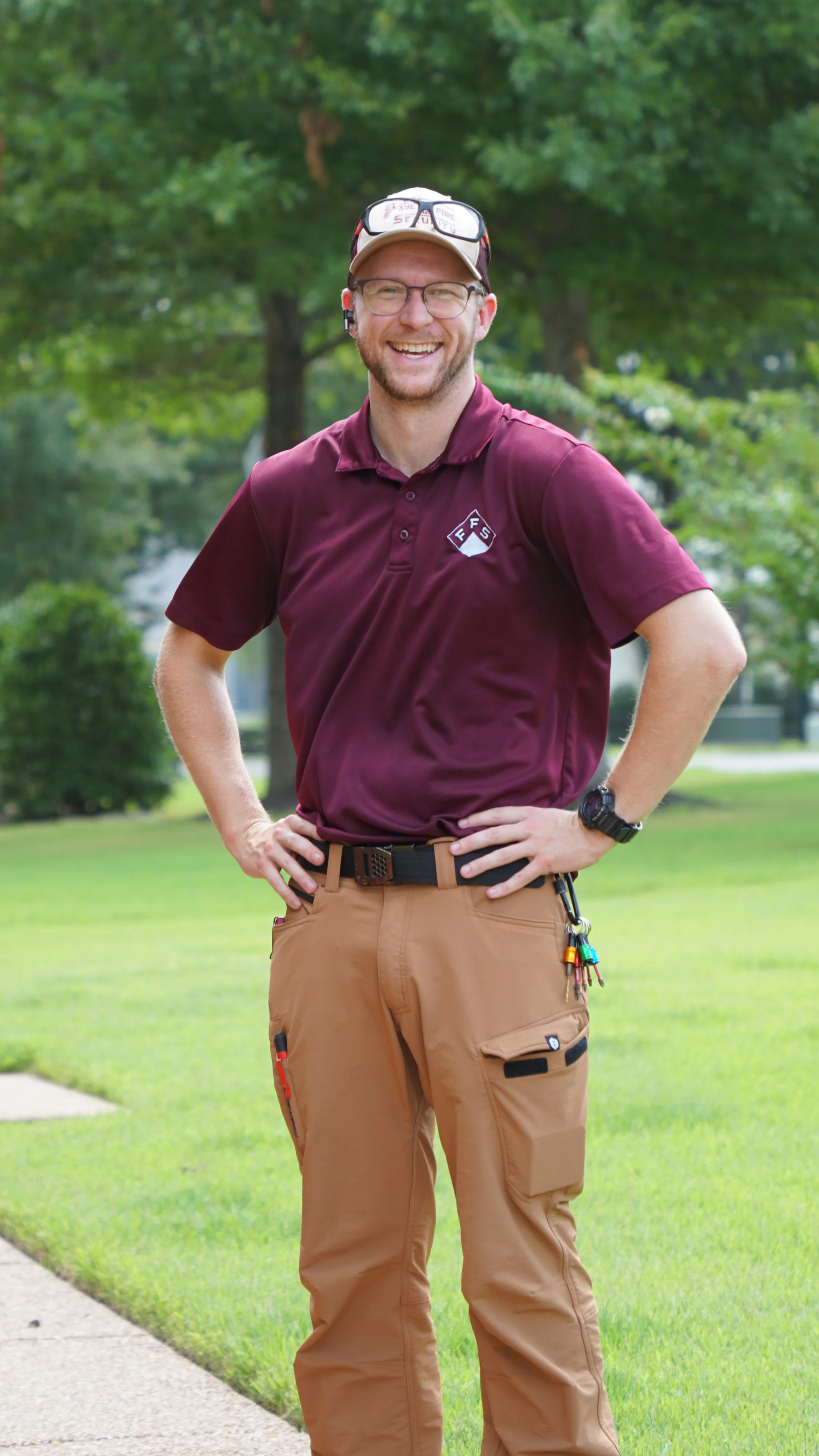 A smiling man wearing safety glasses, a maroon polo shirt with a fire department logo, tan cargo pants, a cap, a watch, and sunglasses on his head, standing outdoors on a grassy area with trees in the background.