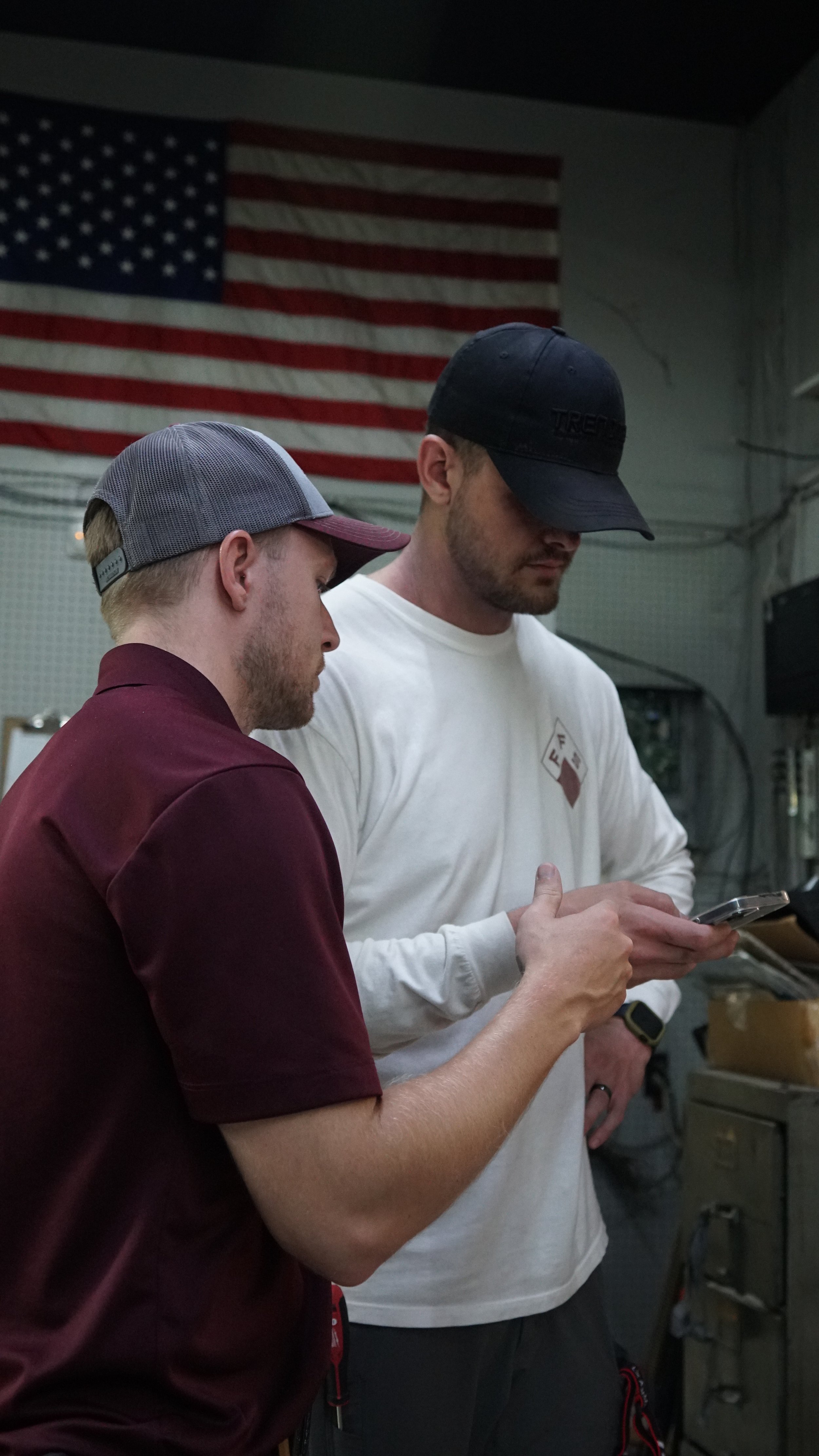 Two men standing close together in an indoor setting, looking at a smartphone, with an American flag hanging on the wall behind them.
