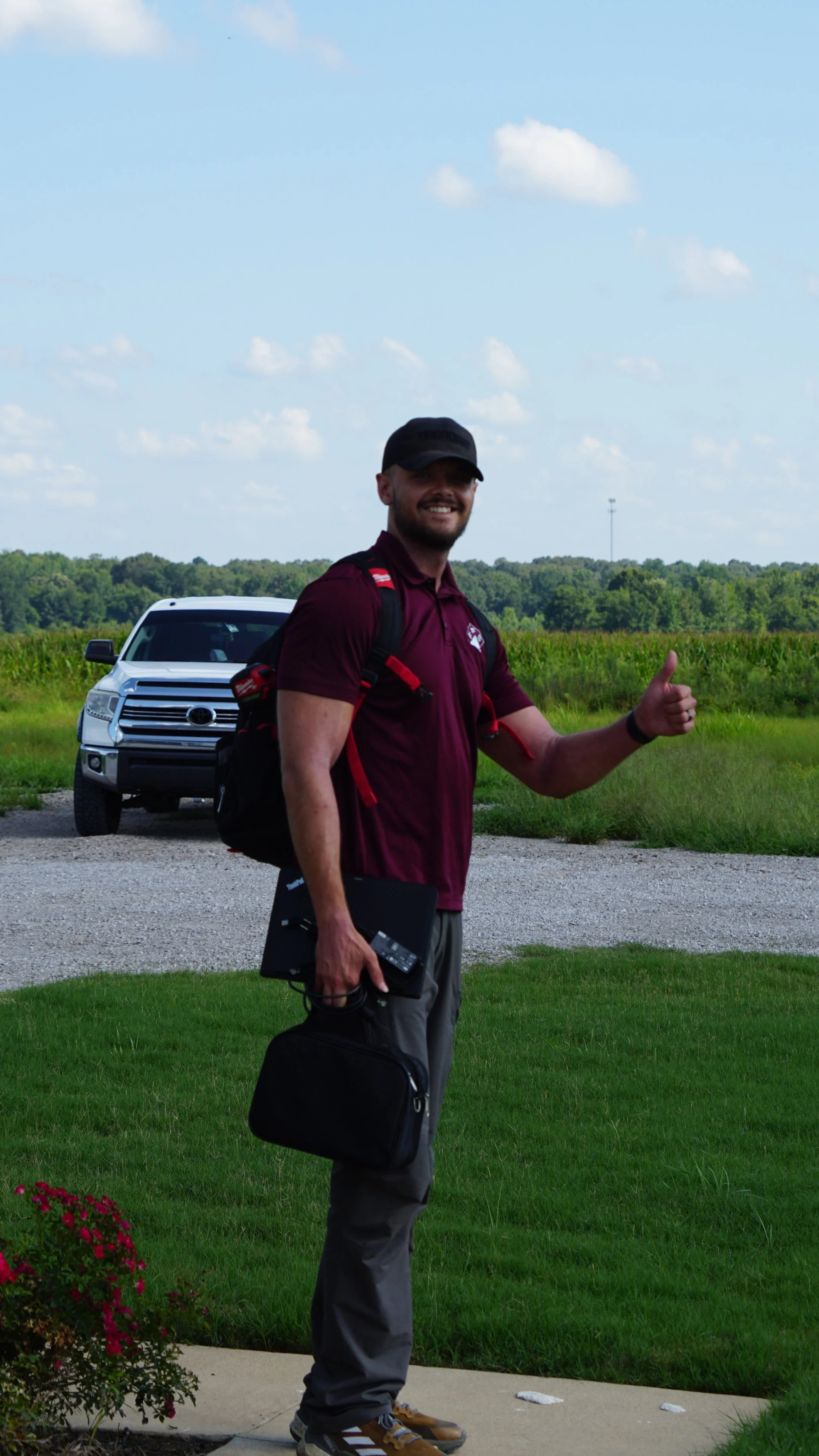 A man in a maroon shirt and a black cap standing on grass with a black case in his hand, giving a thumbs-up, with a silver SUV and open field in the background.