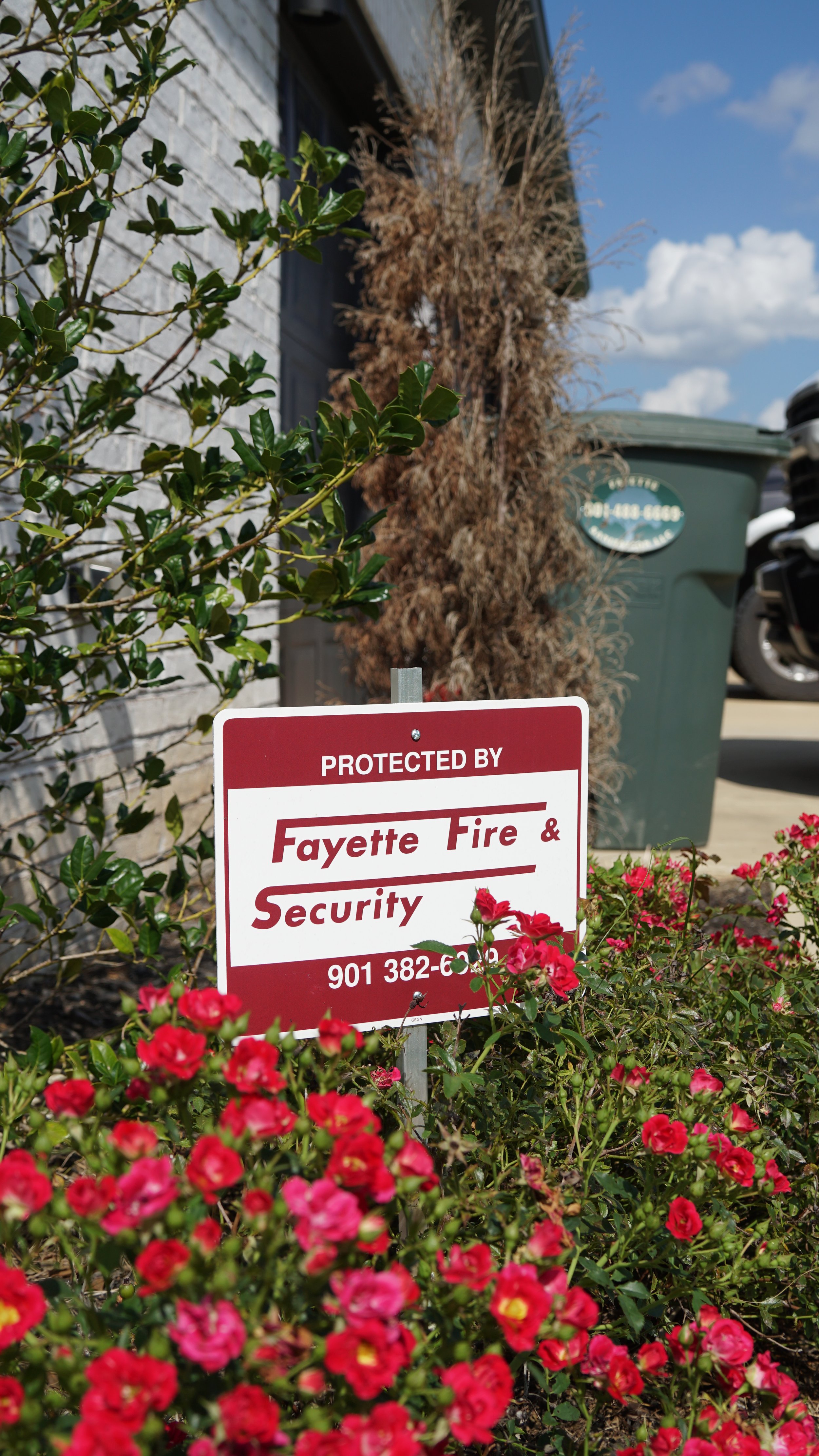 A security sign in front of a garden with pink flowers, a green trash bin, and a brown dried plant in the background.