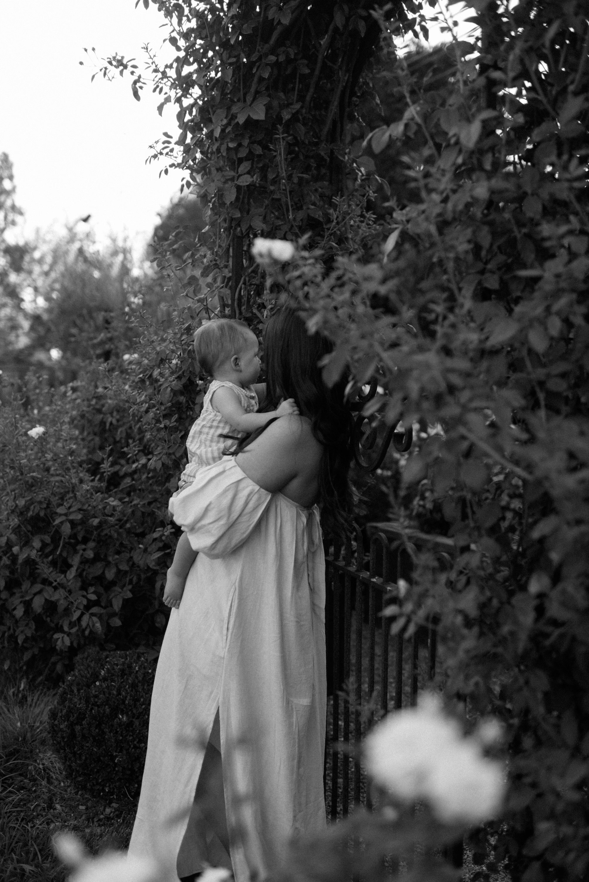 A woman holding a young girl on her shoulder, standing by a black metal gate surrounded by lush foliage in a garden, captured in black and white.