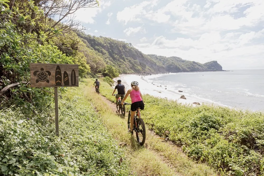 Three people biking on a narrow trail along a lush green coastline with ocean waves, a cloudy sky, and forested hills in the distance.
