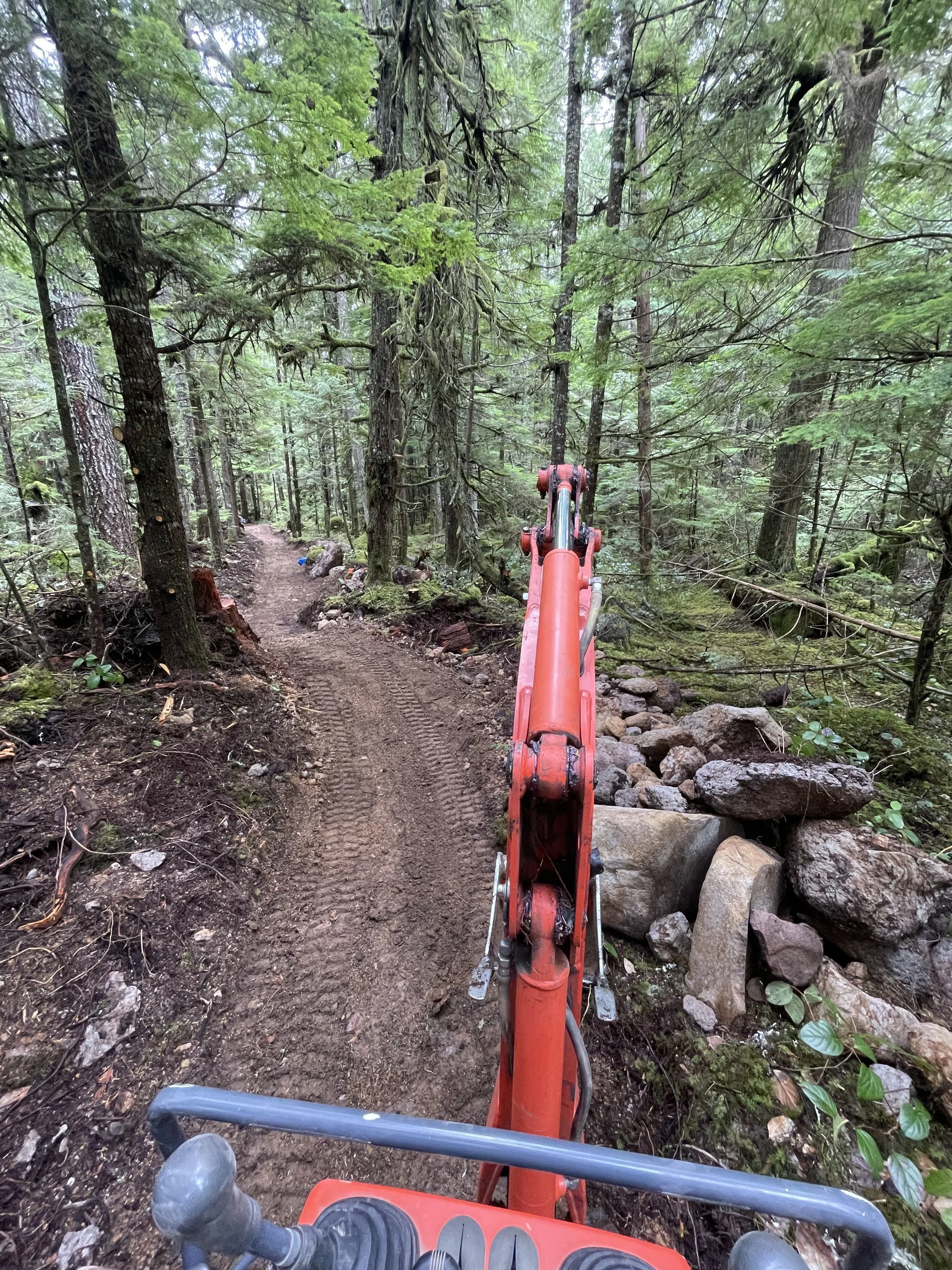 First-person view of a dirt trail through a dense green forest, with the front of an orange excavator or backhoe in the foreground, and trees on both sides of the narrow path.
