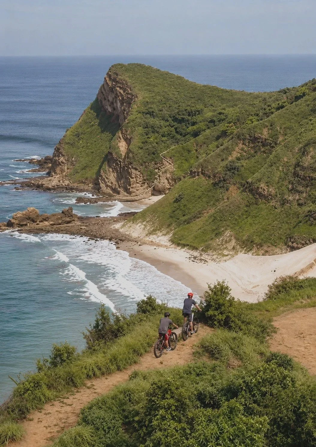 Two people riding bicycles on a dirt trail along a coastal landscape with cliffs, a sandy beach, and the ocean in the background.