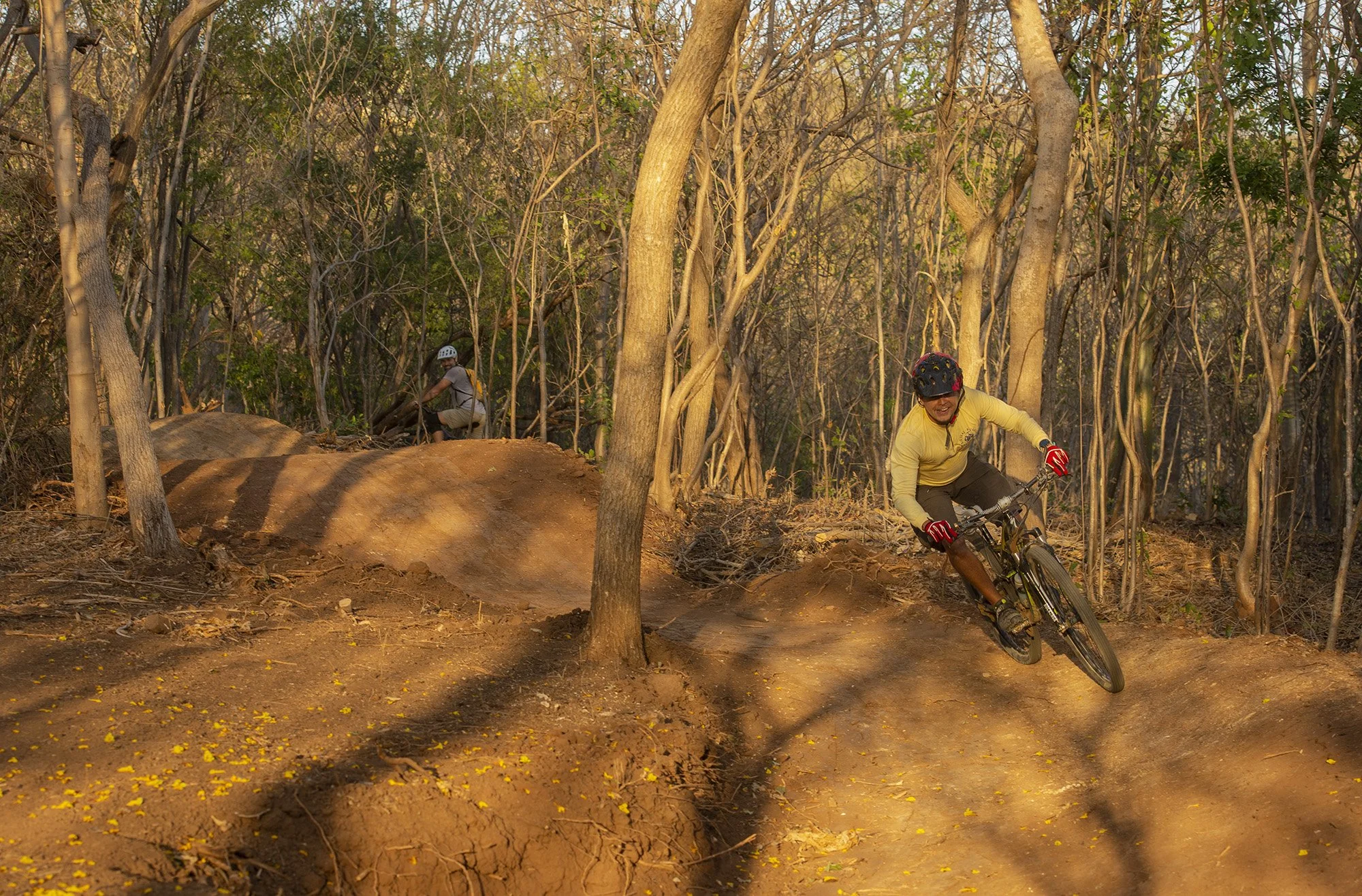 Two mountain bikers riding on a dirt trail through a wooded area, with trees and plants on either side of the path, during daylight.