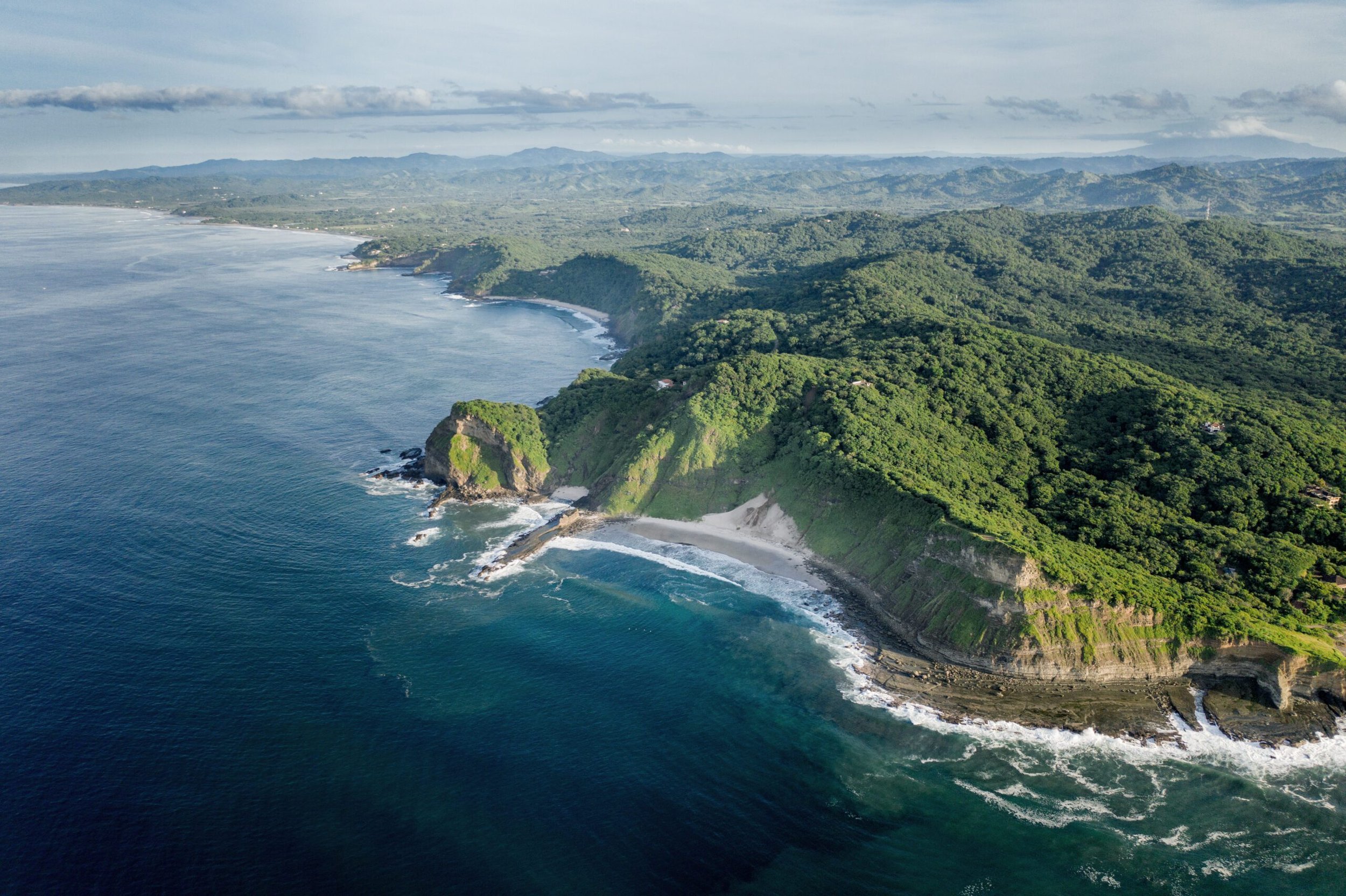 Aerial view of lush green cliffs and coastline meeting the ocean, with waves crashing against the rocks, under a cloudy sky.