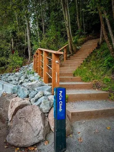 A wooden staircase with a railing leading up a forested trail, next to a sign that reads 'Poc Climb,' and rocks on the ground nearby.