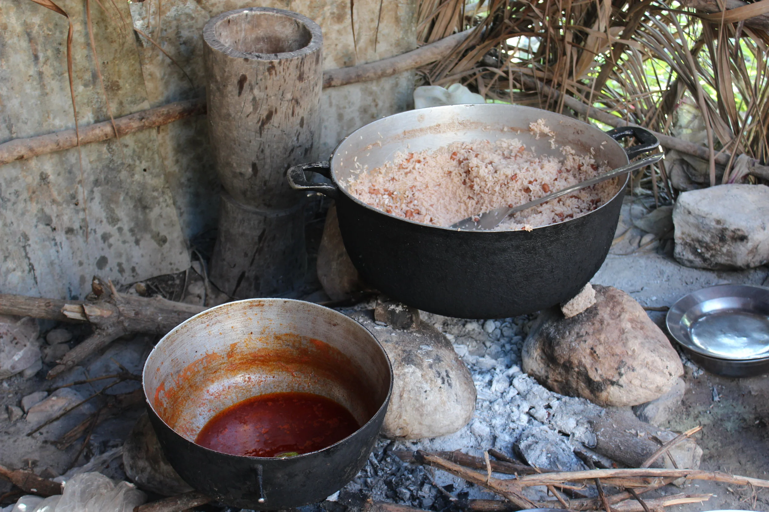A large black pot filled with rice and beans, sitting over an open fire in an outdoor rustic setting with rocks and sticks around.
