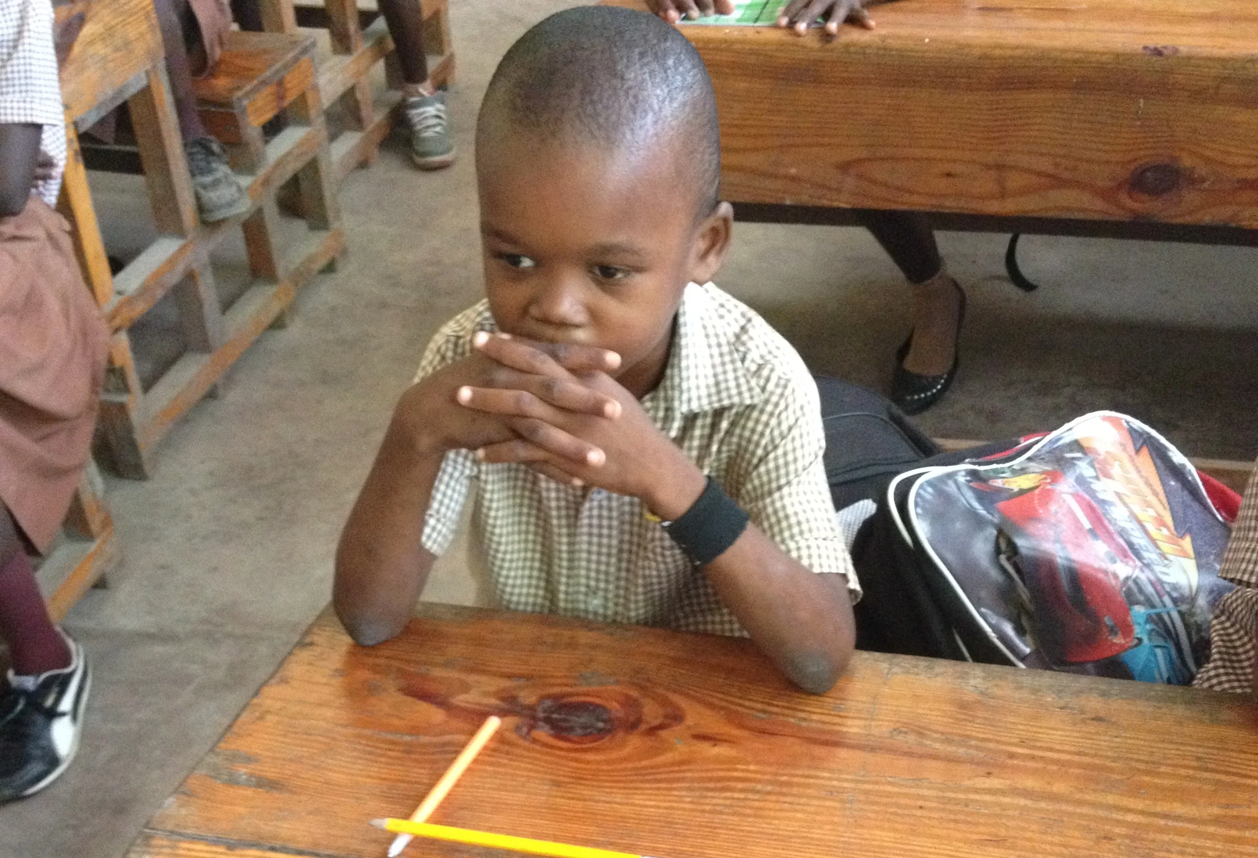 A young boy with short hair sitting at a wooden table, resting his chin on his clasped hands, wearing a checkered shirt and a smartwatch, with a school backpack beside him.