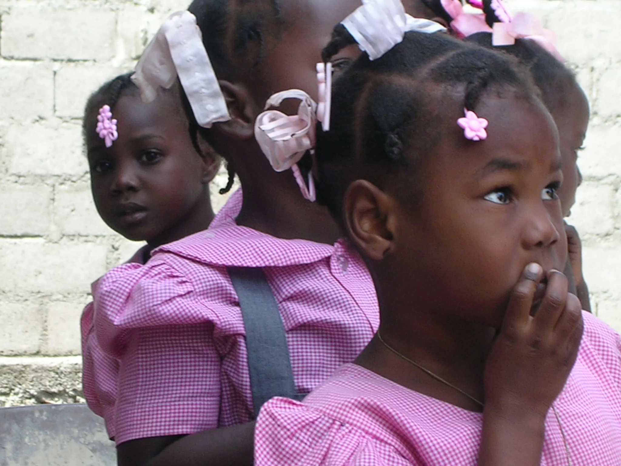Three young Black girls with braided hair, adorned with pink floral hair accessories, wearing pink checkered dresses. They are standing close together outdoors against a light-colored brick wall, engaging in a playful or intimate moment.