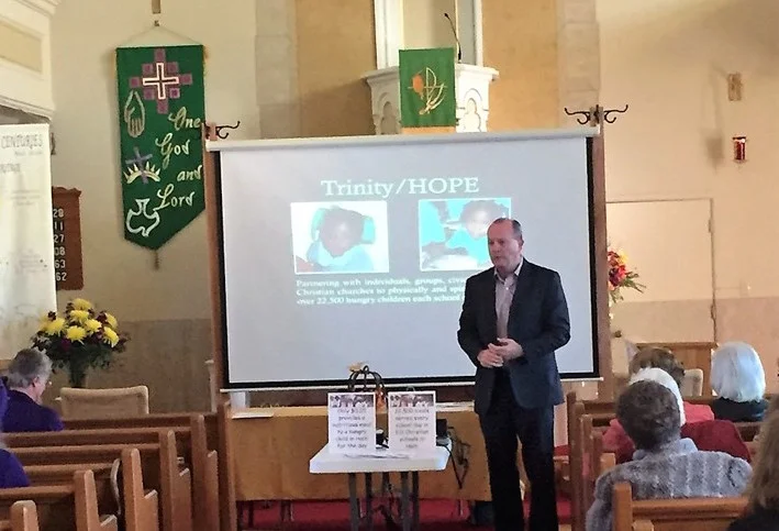 A man in a suit giving a presentation in a church, with a projector screen behind him displaying the words 'Trinity/HOPE' and images of children. The church has green banners with religious symbols, flower arrangements, and wooden pews with an audience of seated adults.