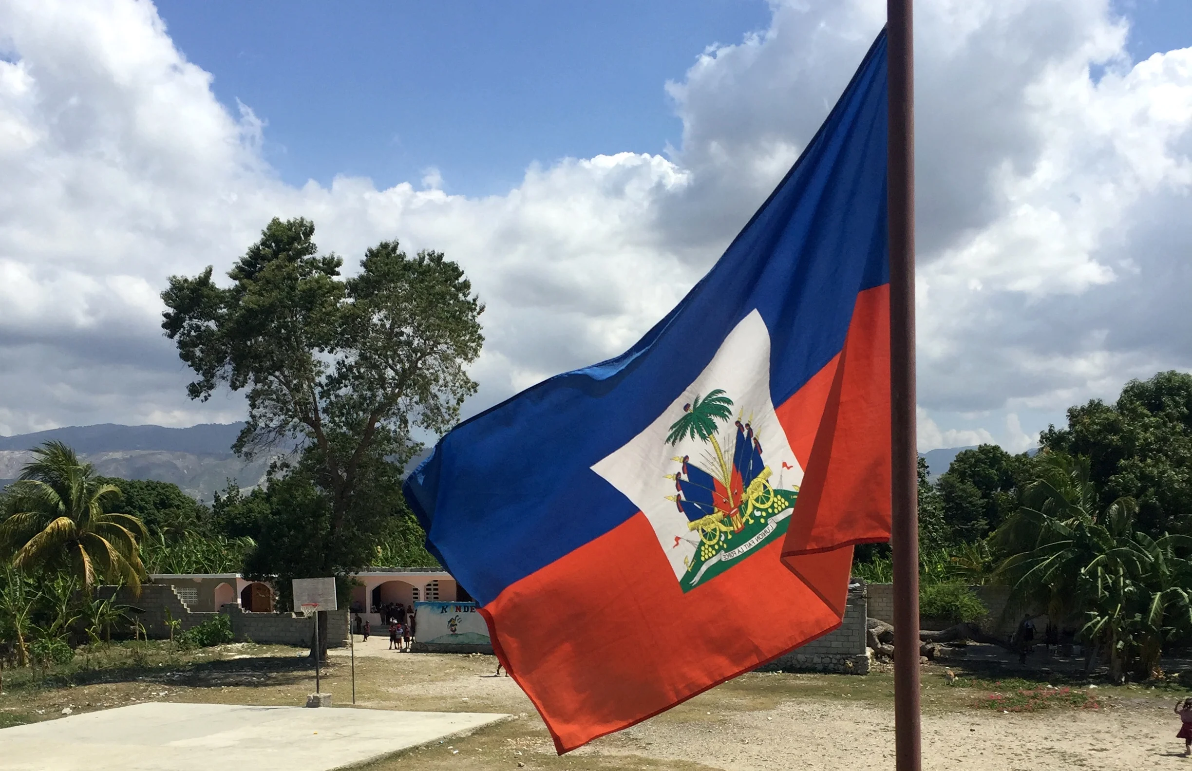 Haitian flag with blue, white, and red stripes and a coat of arms in the center, flying outdoors under a partly cloudy sky with trees and mountains in the background.