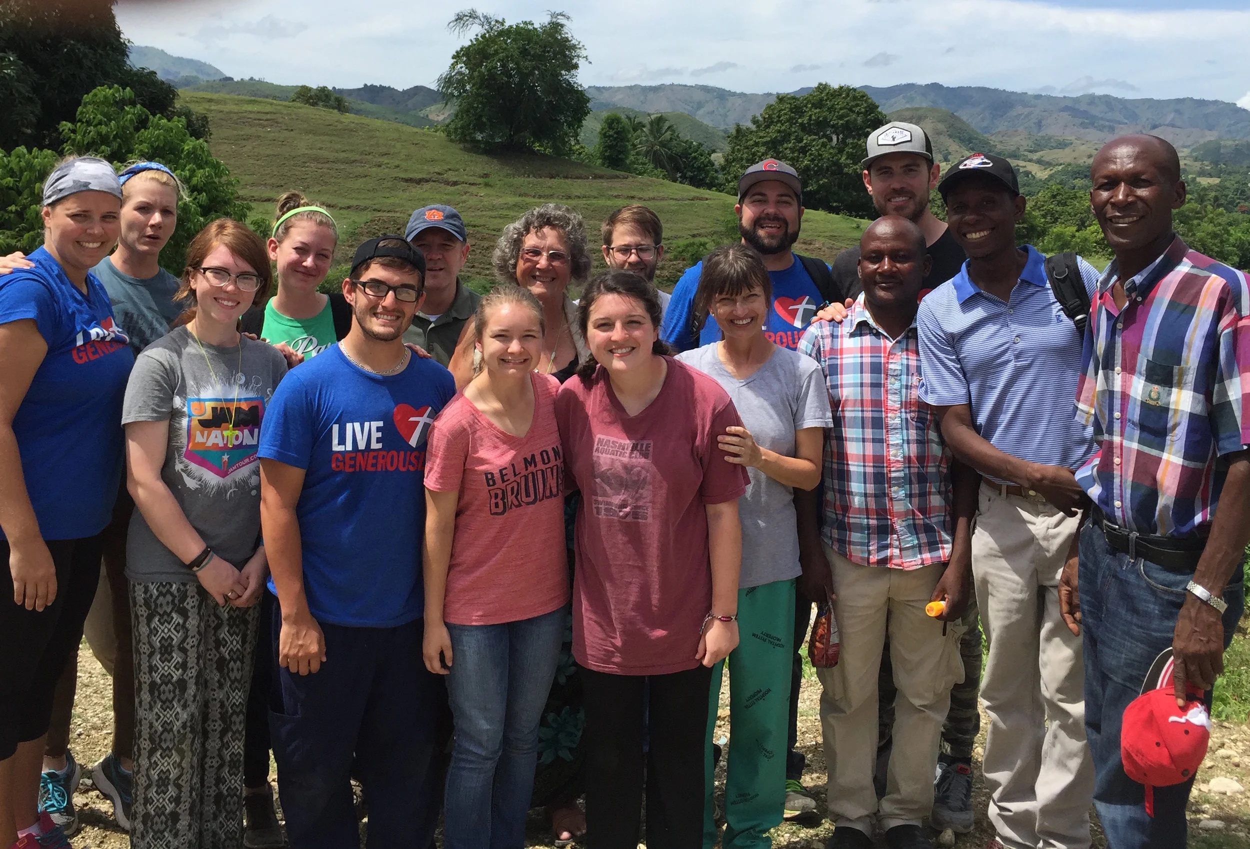 Group of people outdoors in a hilly, green landscape with trees and mountains in the background.