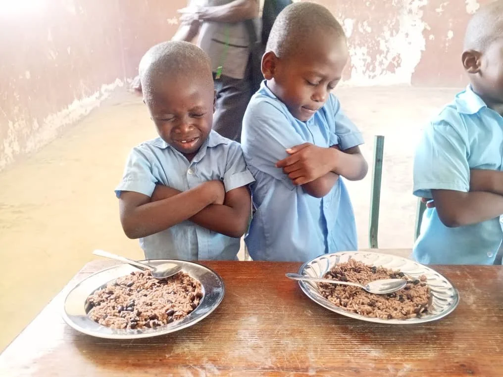 Two children in blue school uniforms with arms crossed, looking displeased at plates of food on a wooden table. The food appears to be rice and beans.