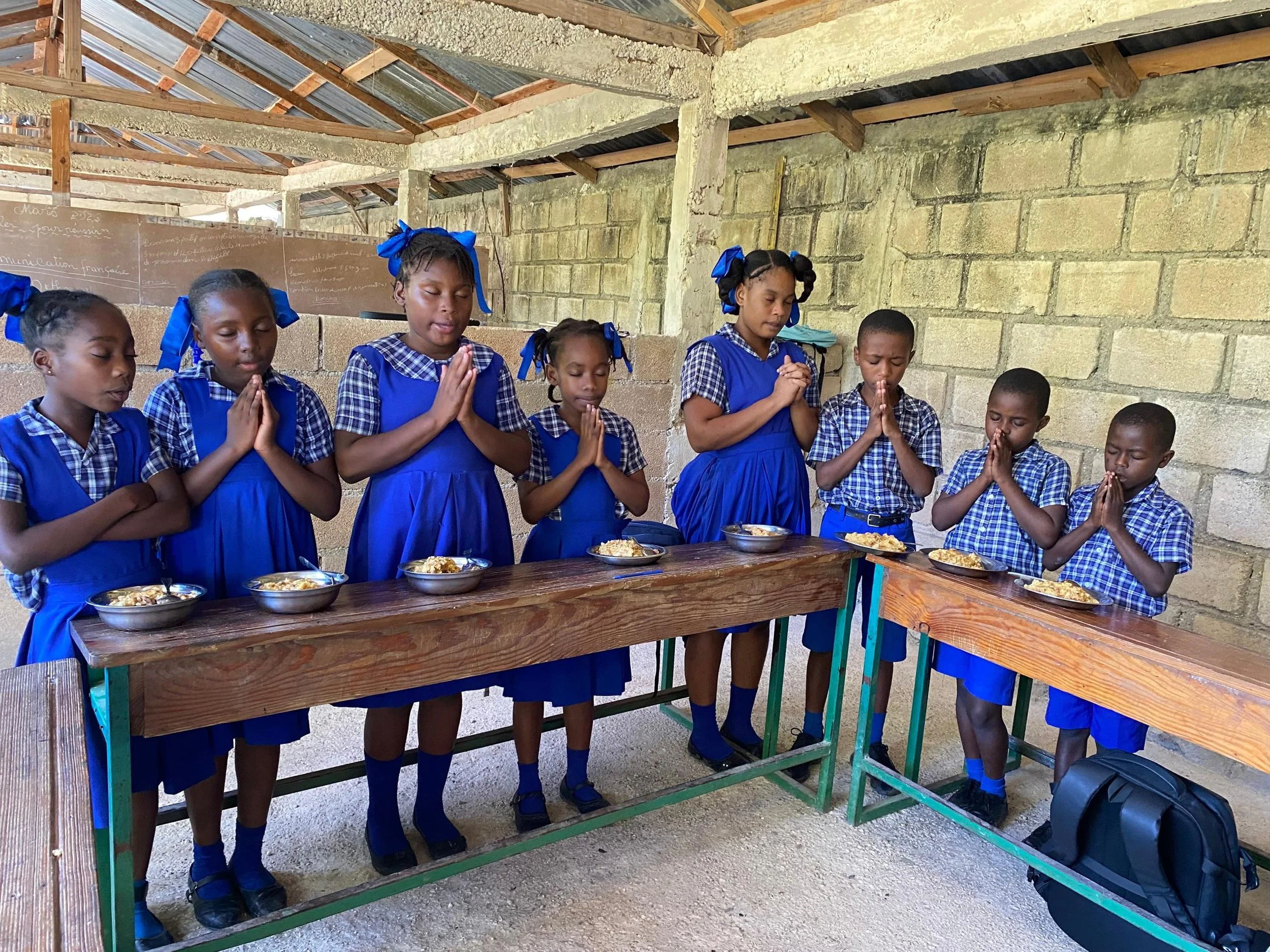 Young girls dressed in blue school uniforms and bows, praying with eyes closed and hands together, standing behind a table with bowls of food inside a rustic, unfinished building.