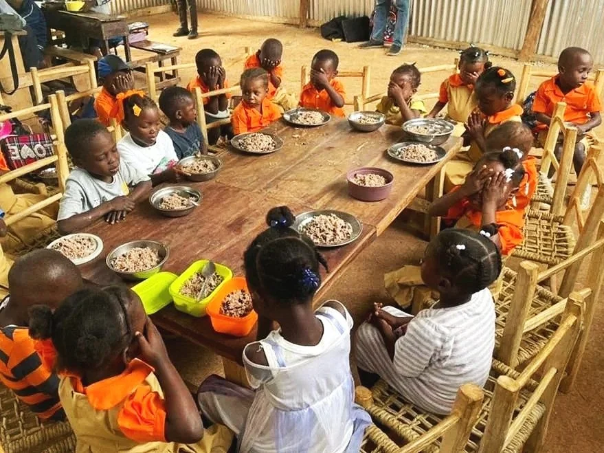 Children sitting around a large wooden table, eating a meal from metal and plastic bowls, in a room with a dirt floor and corrugated metal walls.