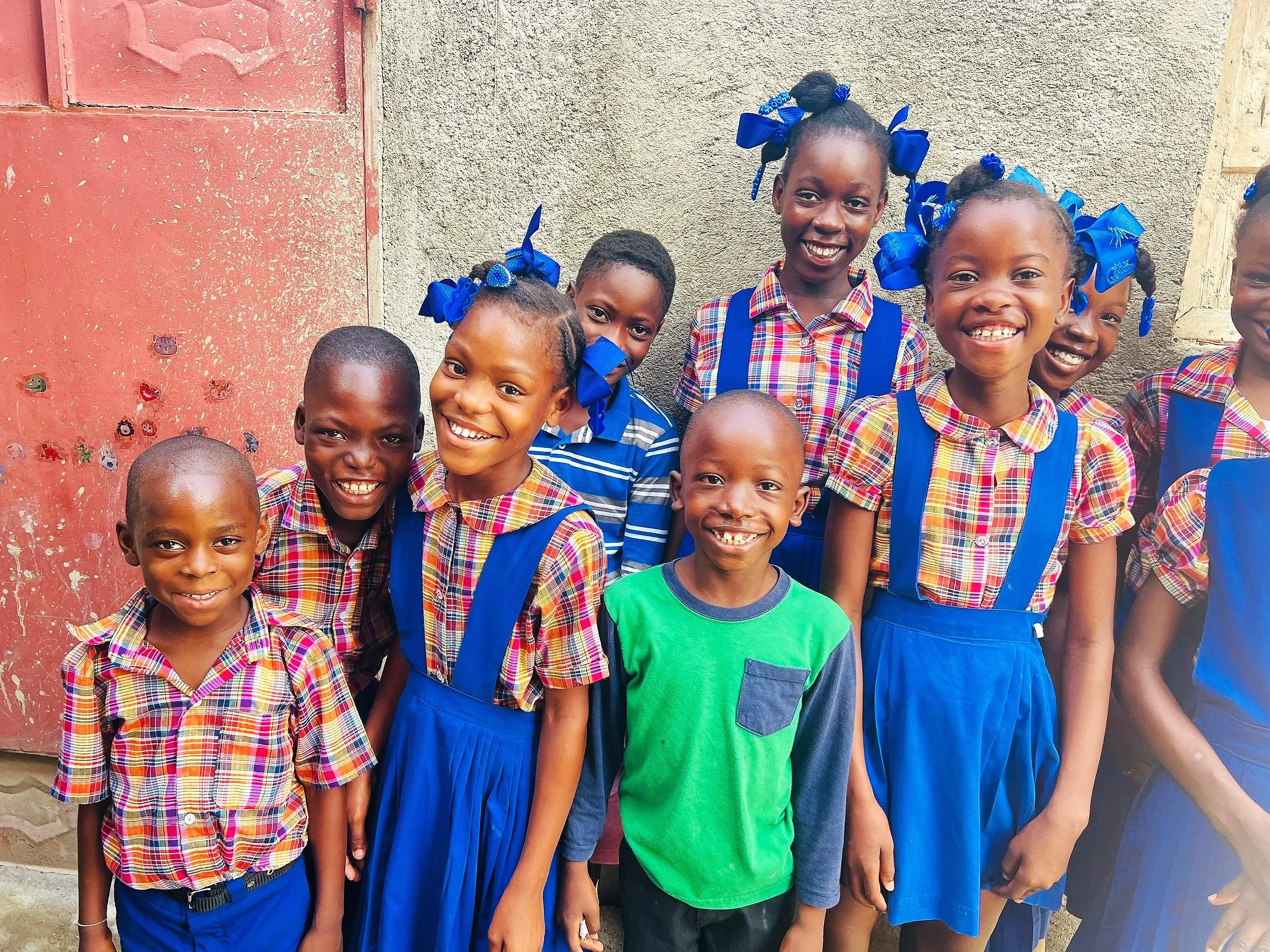 A group of happy children, mostly girls, dressed in matching colorful plaid uniforms with blue suspenders, standing in front of a textured wall, smiling at the camera. Some girls have blue bows in their hair.