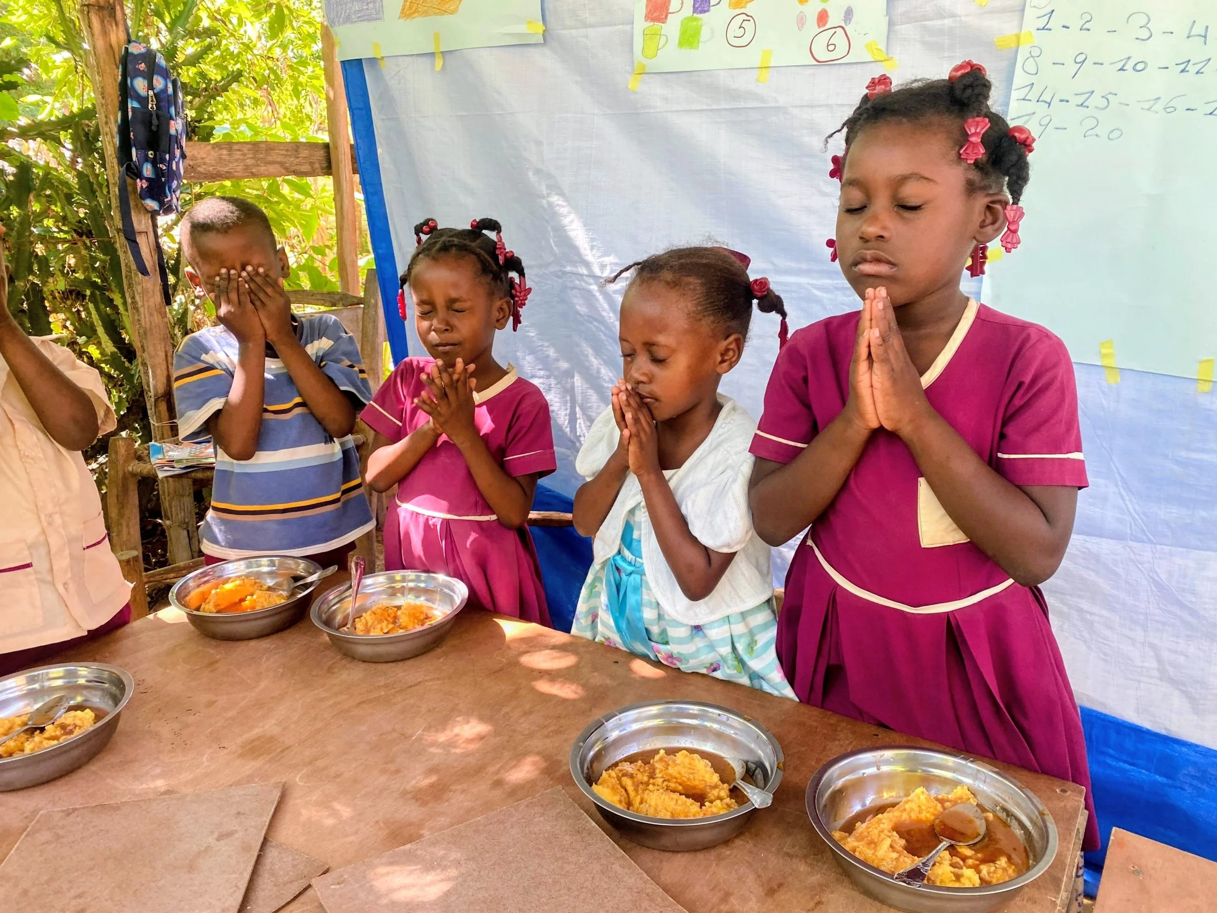 Group of young children praying around a table with bowls of food, outdoors with trees and educational posters in the background.