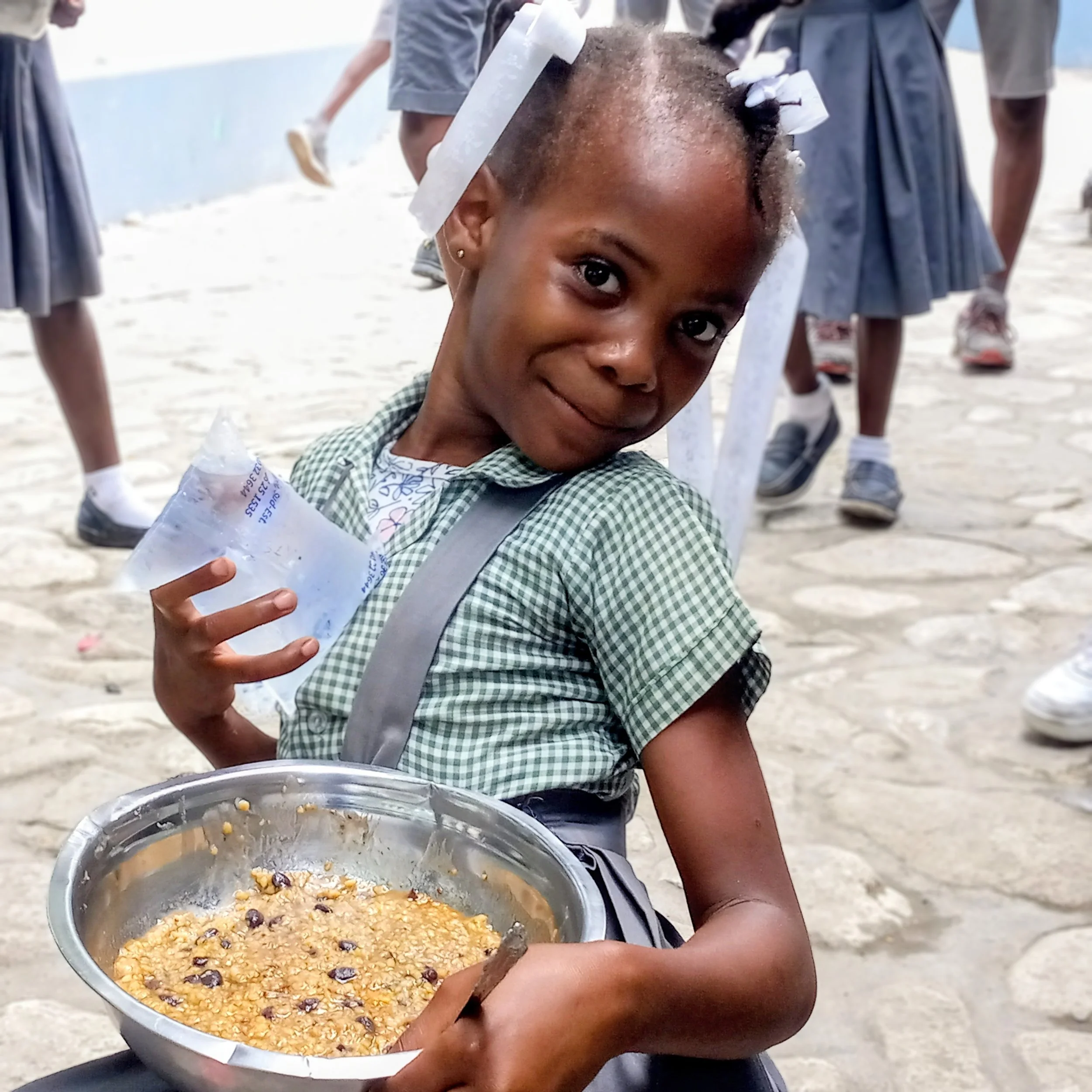 Child sitting outdoors holding a metal bowl filled with cooked rice and beans, wearing a green checkered shirt, with other children in school uniforms in the background.