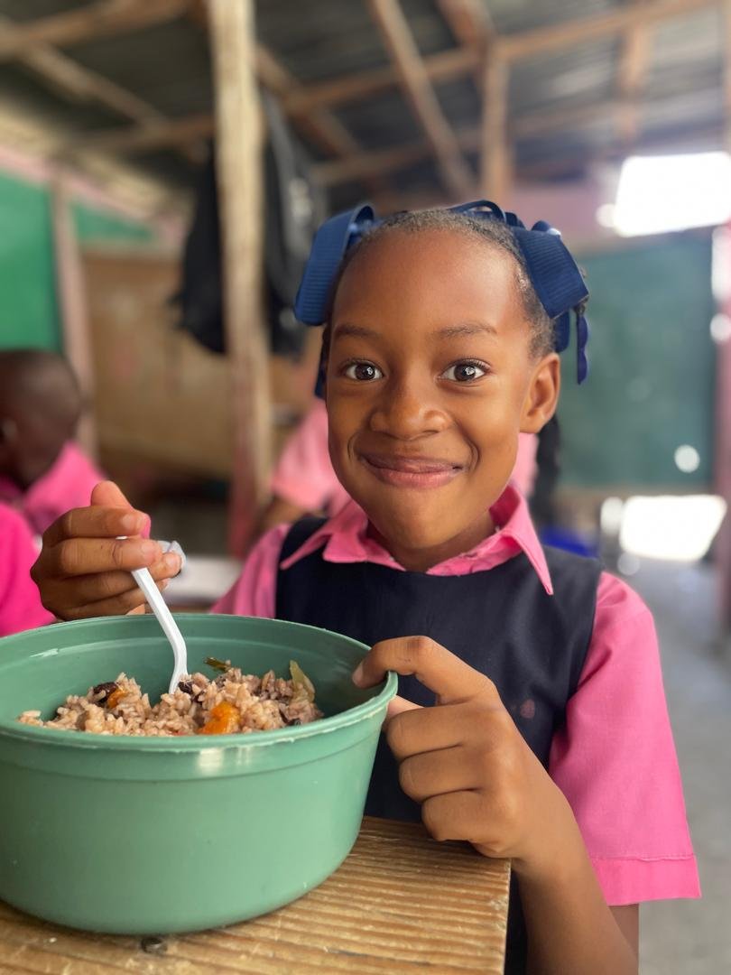 A smiling young girl in pink and navy school uniform with a blue hair bow holding a green bowl of rice and vegetables, sitting at a wooden desk in a classroom.