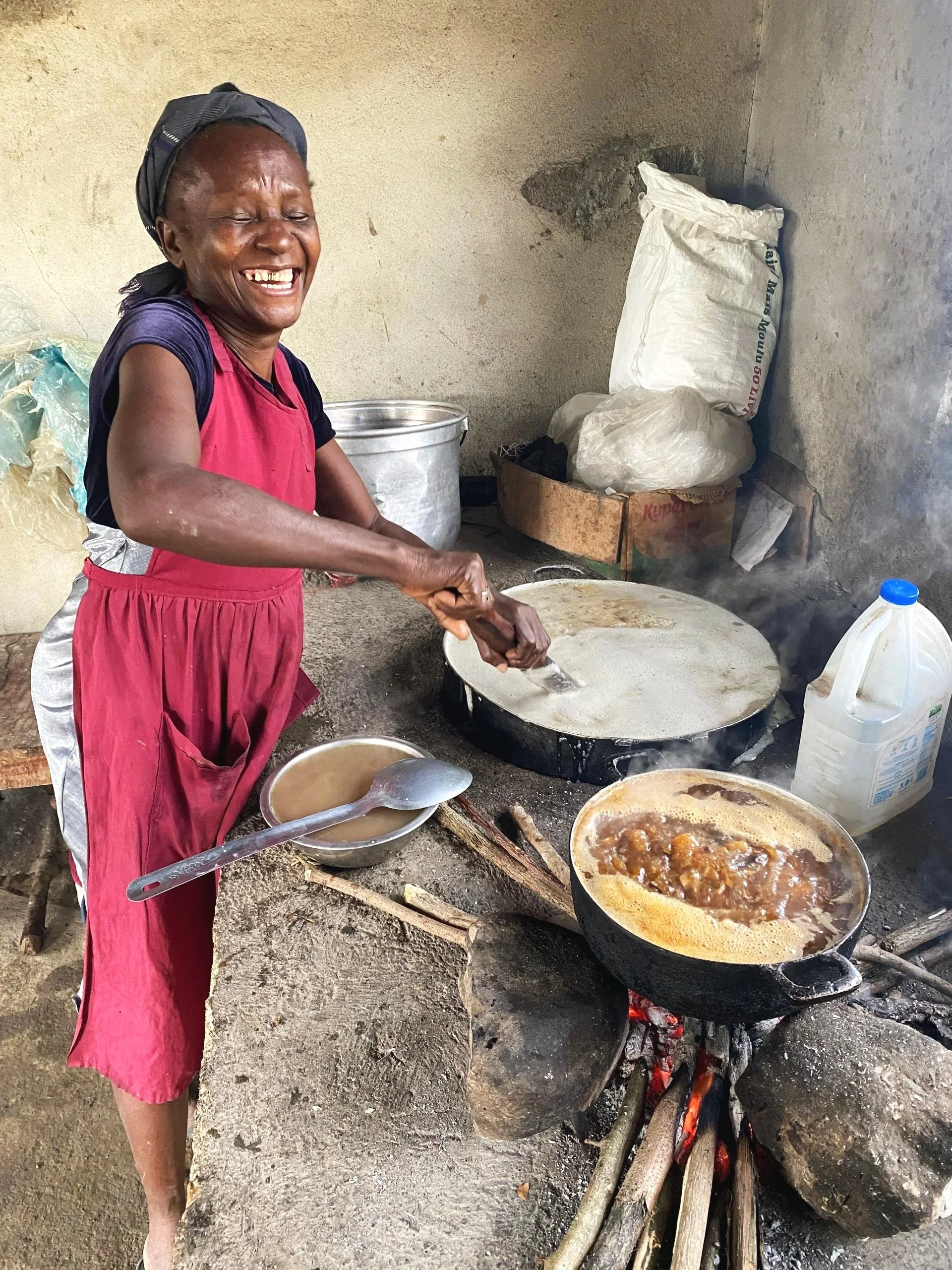 A woman cooking over an open fire in a rustic kitchen, stirring a large skillet with bubbling food, smiling happily.