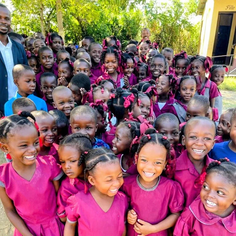 A large group of smiling young girls and a few adults outdoors, wearing matching pink and purple school uniforms with braids adorned with pink ribbons.