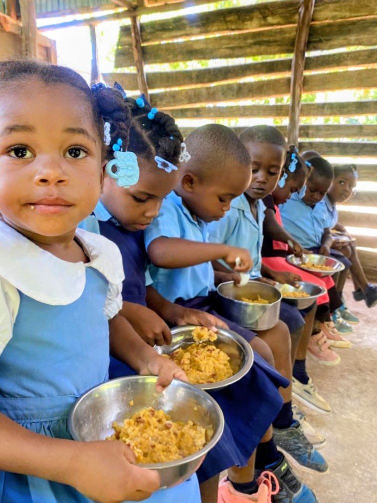 Young children sitting in a row inside a wooden structure, eating from metal bowls, with some looking down at their food and others looking around, wearing school uniforms and sneakers.