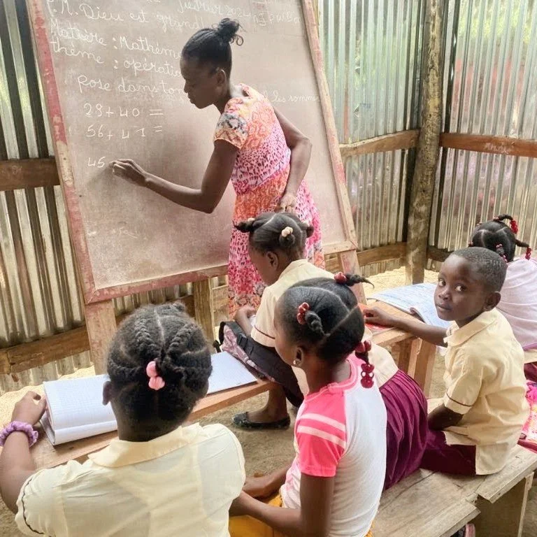 A classroom scene with a teacher at a chalkboard and four young students sitting at desks. The teacher is writing on the blackboard, while the students look on, some taking notes.