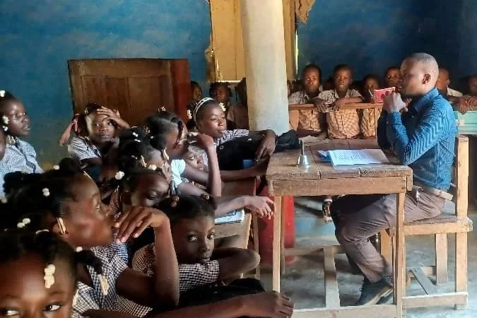 A classroom with young students sitting at desks, listening to a teacher seated at the front.