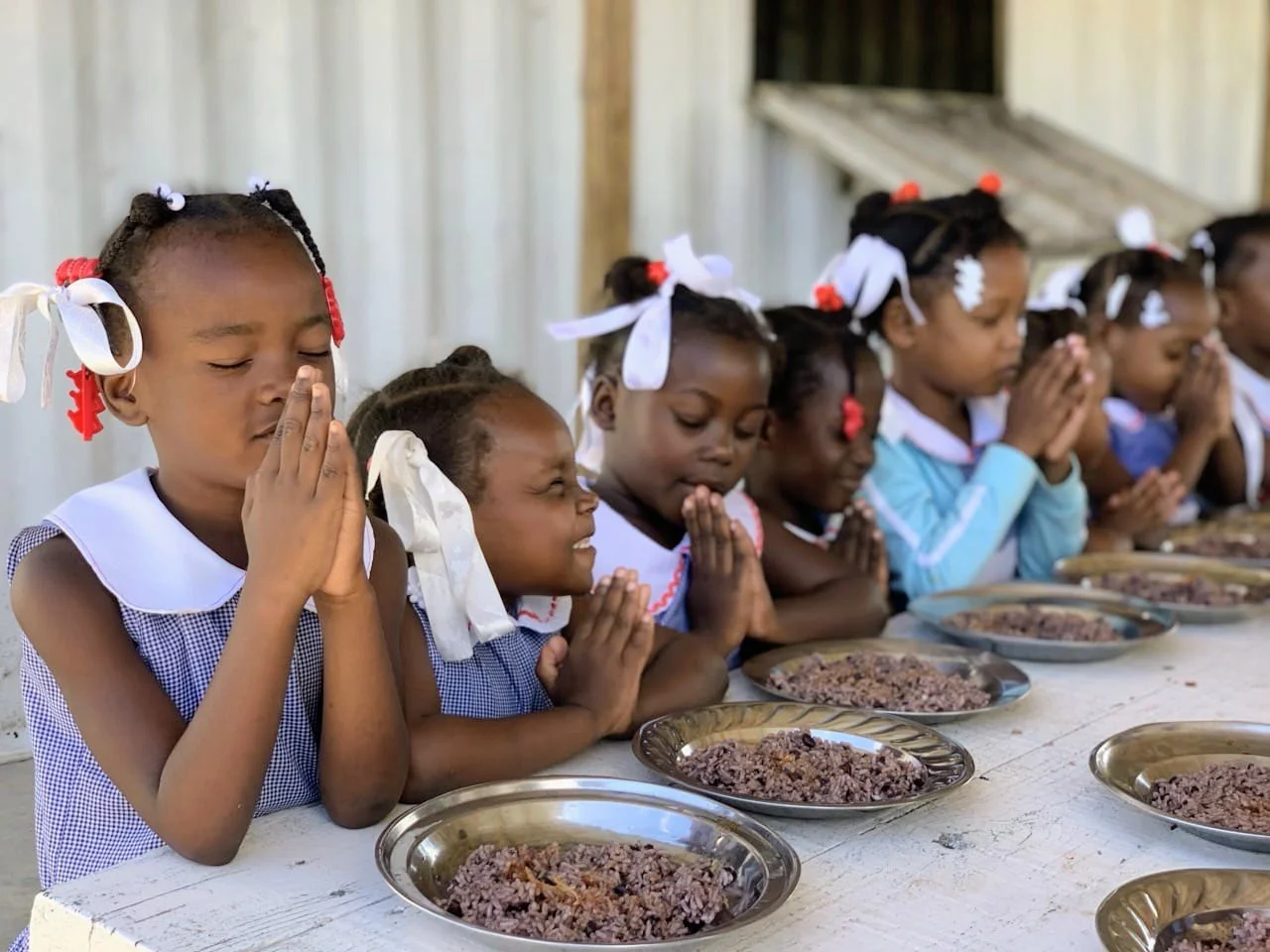 A group of young girls praying before meals at a table with plates of rice, dressed in school uniforms with ribbons in their hair.