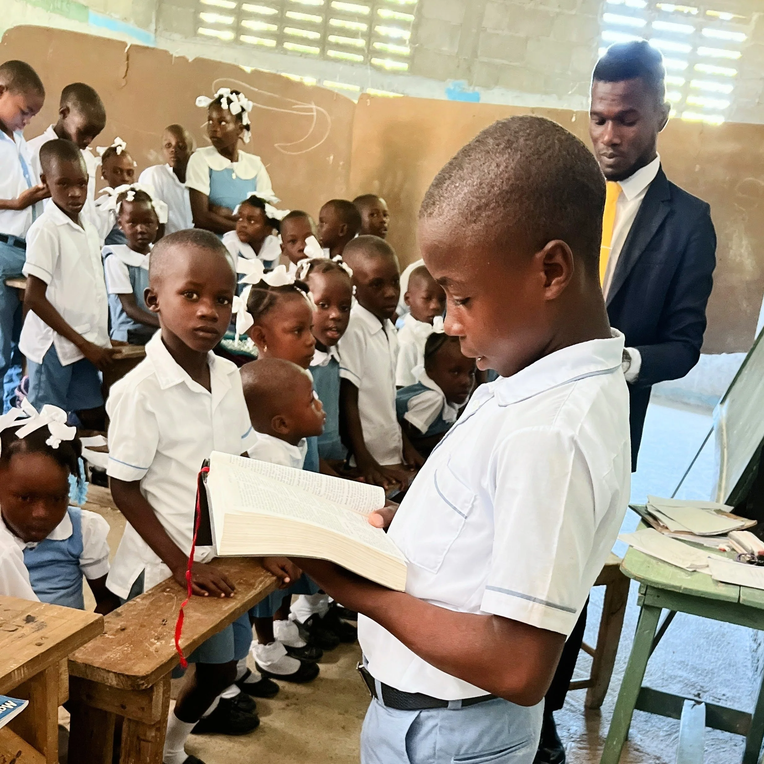 A classroom scene with children wearing uniforms, some standing at desks, a man in a suit and yellow tie looking at a book, and a woman standing nearby. The classroom has a high, partially open ceiling with sunlight coming in.