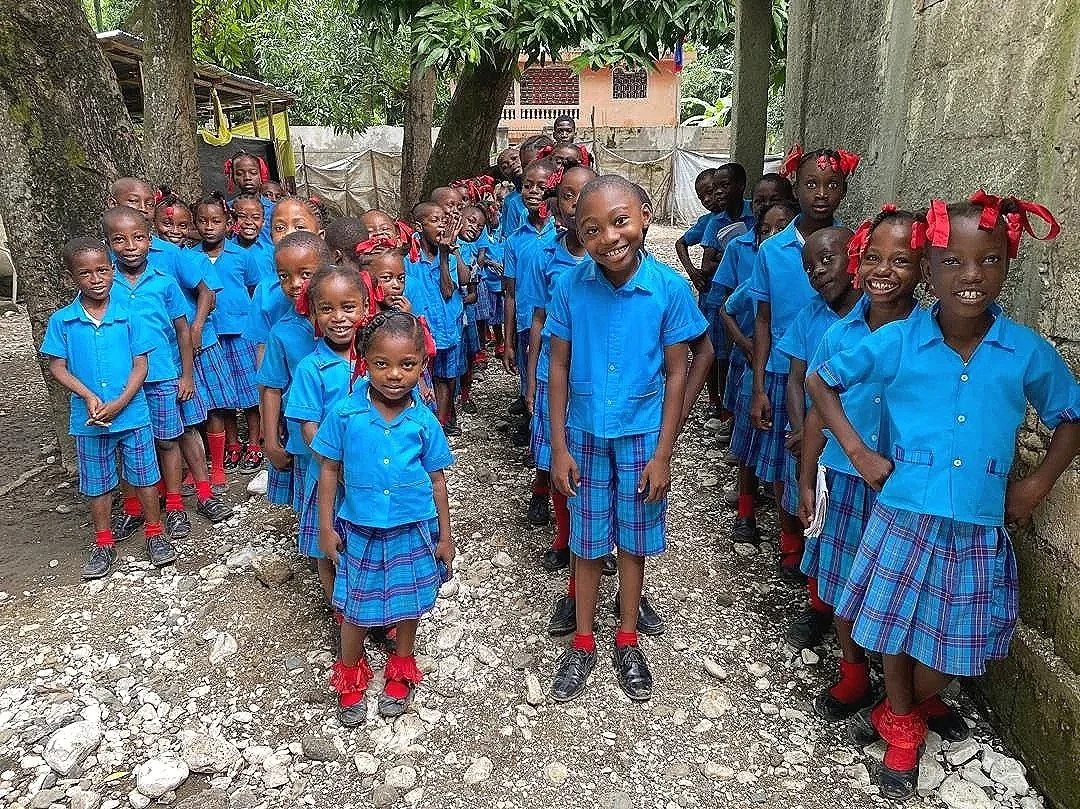 A group of smiling young children in matching blue school uniforms, standing outdoors on a rocky path, with trees and a building in the background.