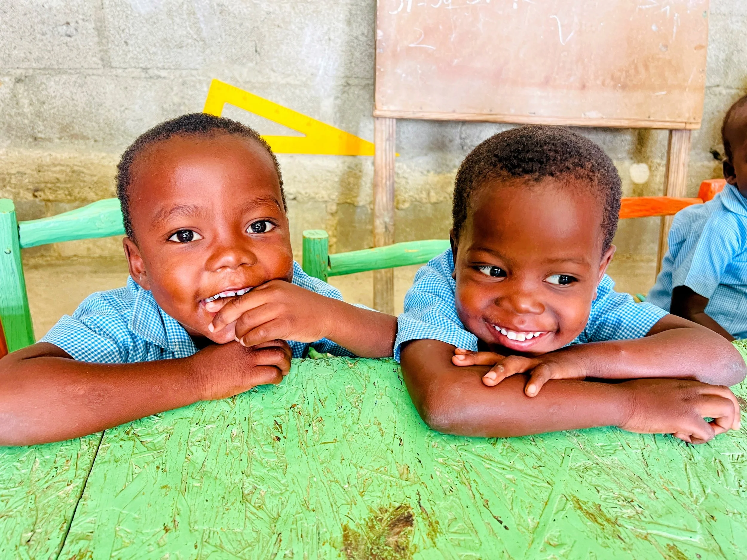 Two young children with short hair and dark skin are smiling and lying on a green wooden table, resting their arms on it, in a classroom setting with a concrete wall and a pink chalkboard in the background.