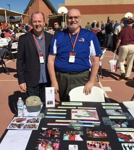 Two men standing behind a table at an outdoor event, with one man wearing a blue sports shirt and the other in a suit. The table displays various flyers, photos, and a water bottle, with a crowd in the background.
