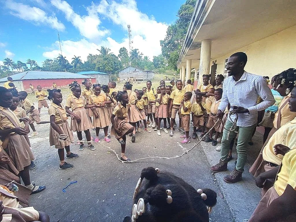 A group of school children in yellow uniforms are gathered outside, watching a man jump rope with two young children. The scene takes place outdoors on a sunny day.
