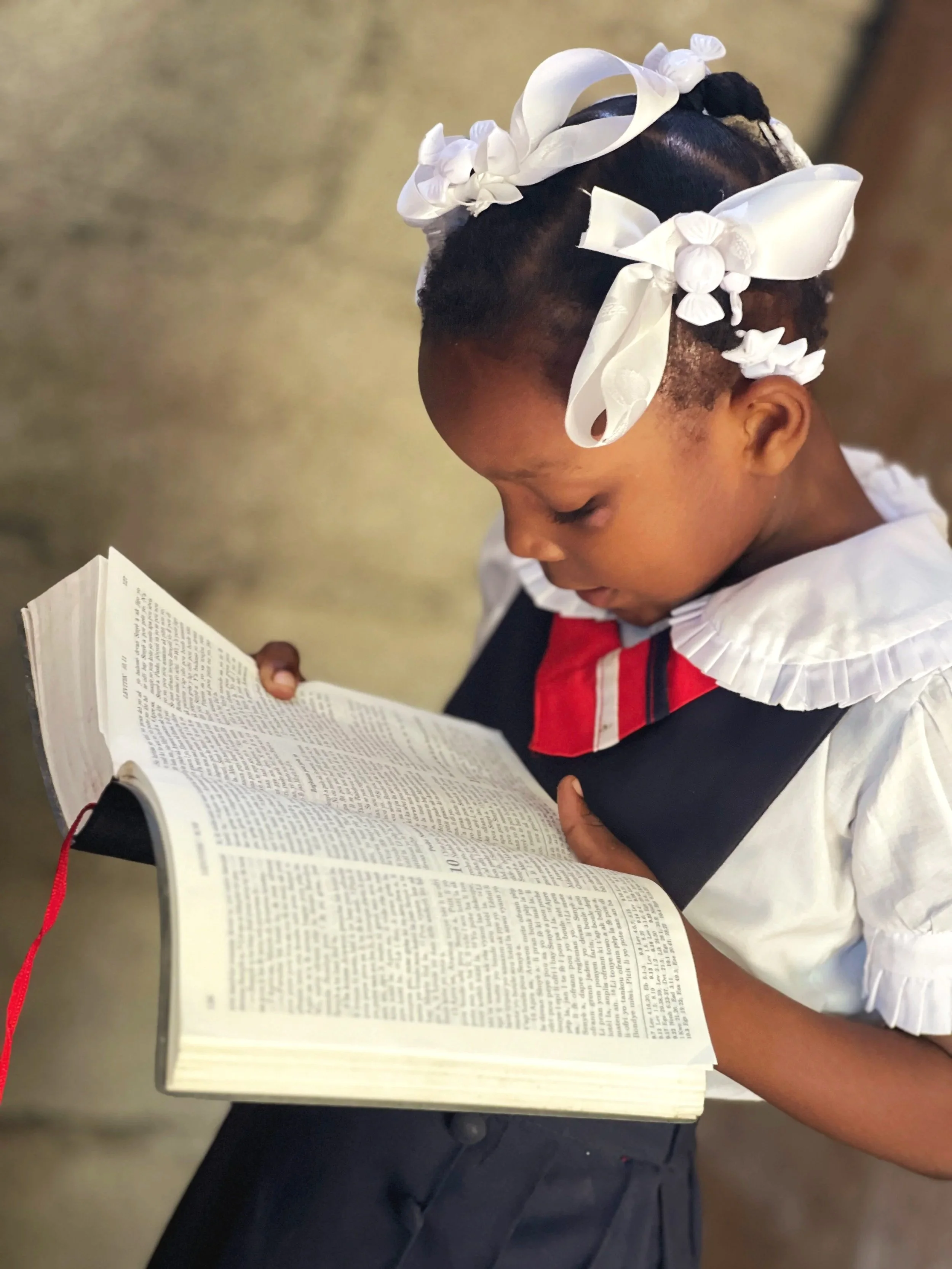 A young girl with dark skin and short hair, decorated with white ribbon bows, is reading a white book with small text. She is wearing a white shirt with a red, black, and white striped tie.