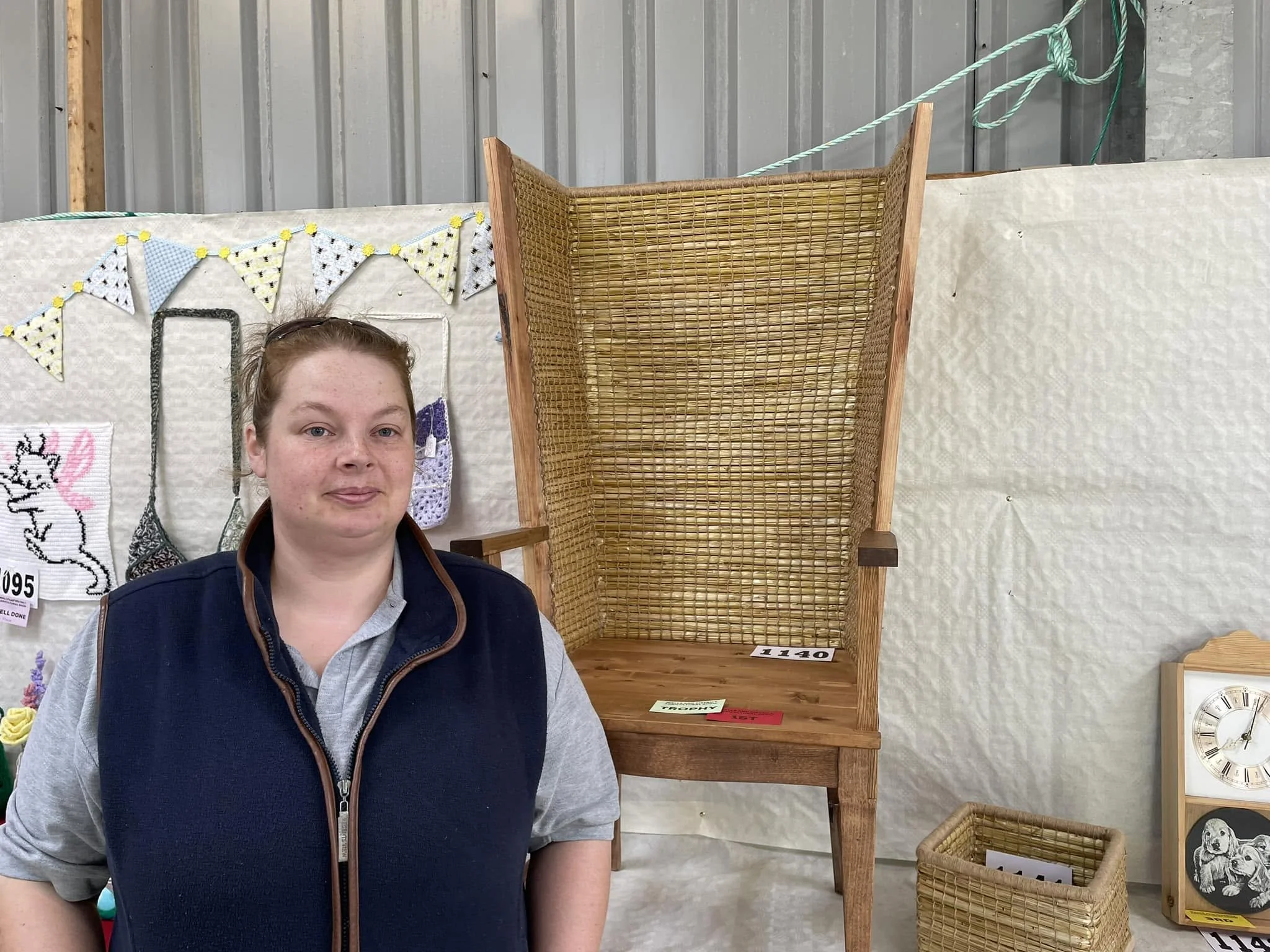 A woman with reddish hair and a vest standing in front of a display of handmade crafts, including baskets, cross-stitch art, and a wall clock, at a county show.
