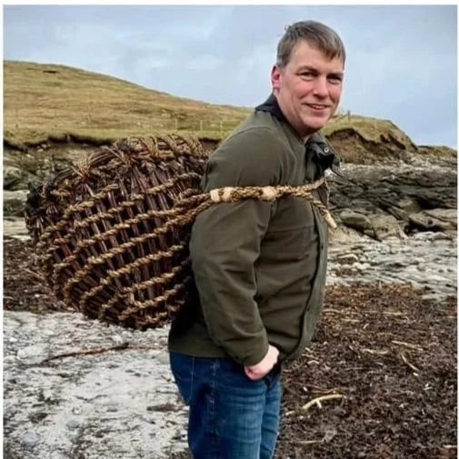 Man standing on rocky shoreline with a basket strapped to his back, near grassy cliffs and the ocean in the background.
