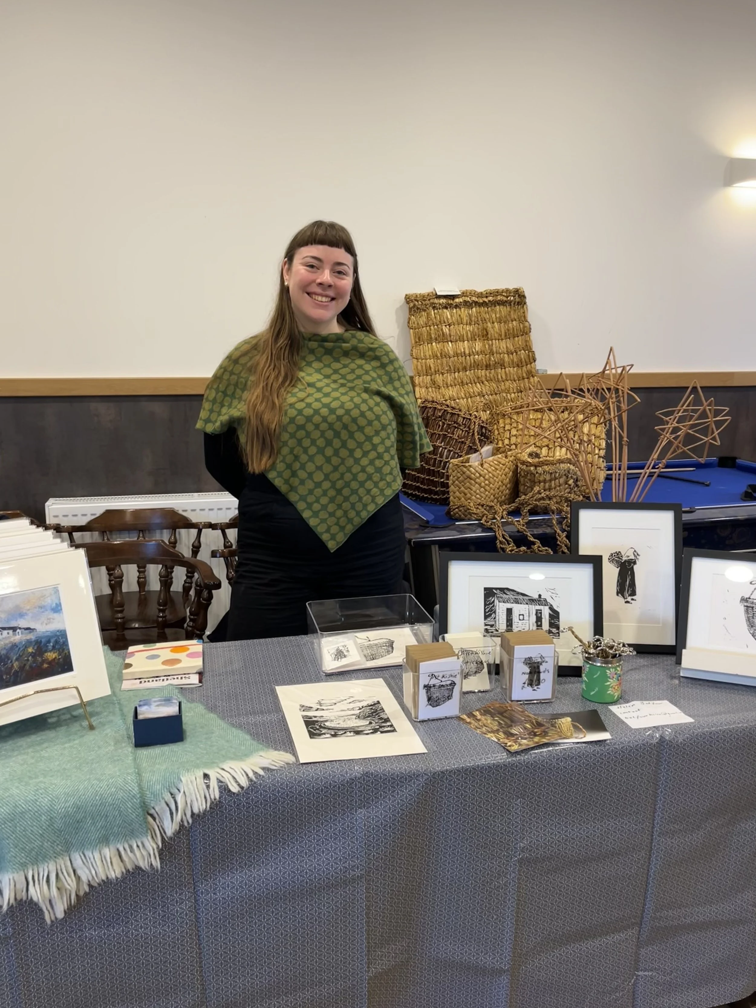 A woman smiling at a craft fair, standing behind a table displaying artwork, handmade woven baskets, and decorative items. There are framed pictures, jewelry, and small craft items on the table.