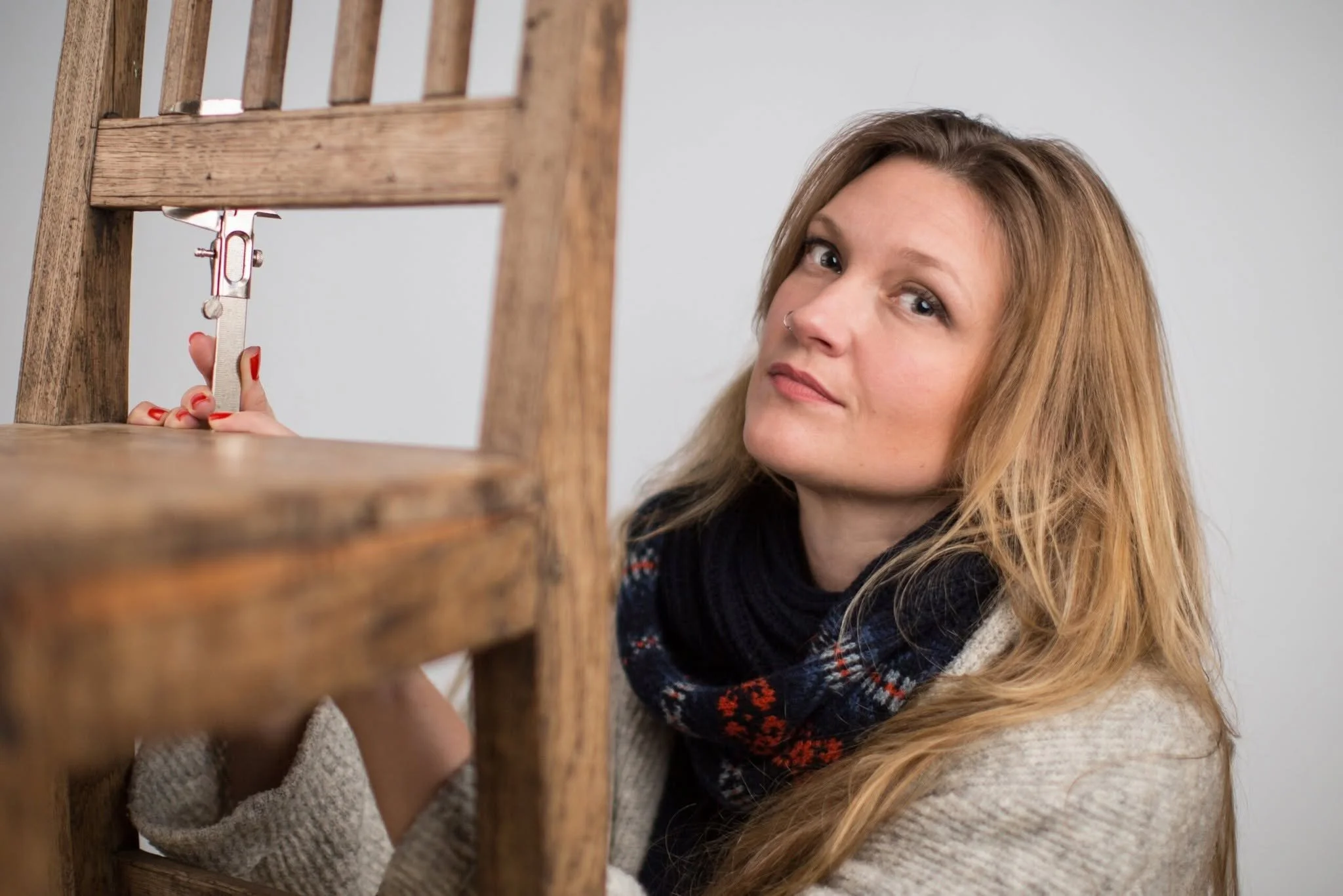 A woman with long, wavy hair and a nose ring measures a wooden furniture piece with a ruler.
