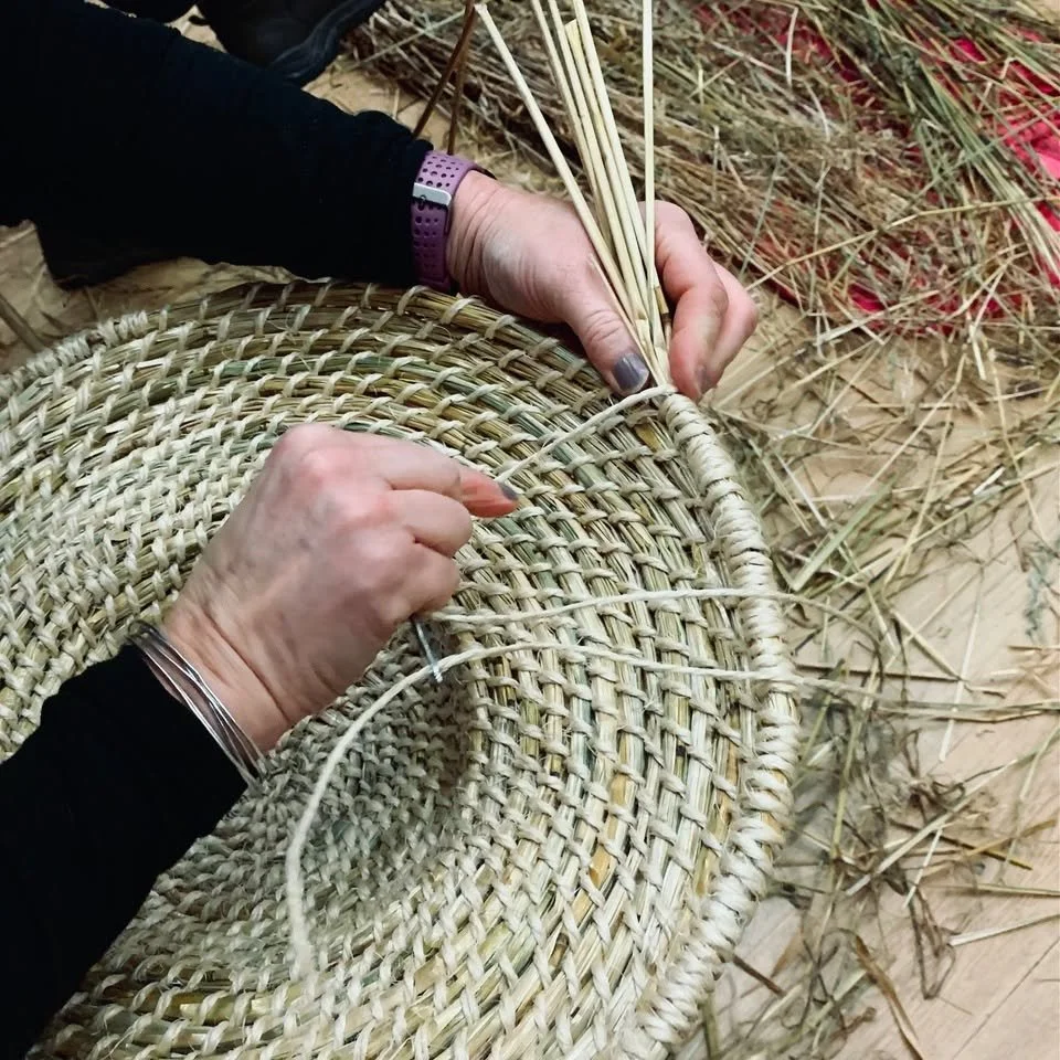 A person weaving a basket with straw, surrounded by straw and weaving materials.