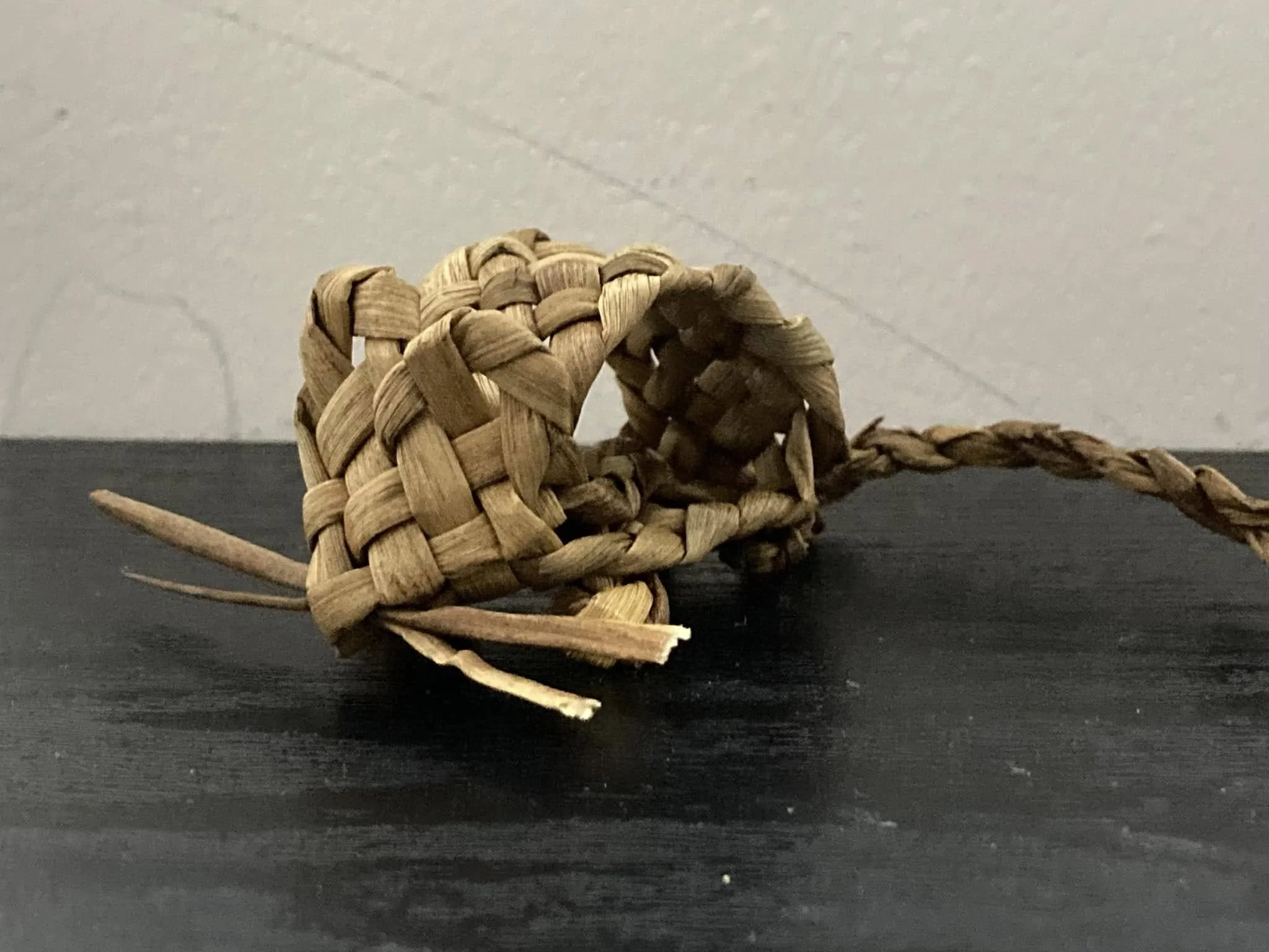 A small woven decorative mouse made of soft rush, with a round body, a tail, and a head, placed on a black surface against a grey wall background.