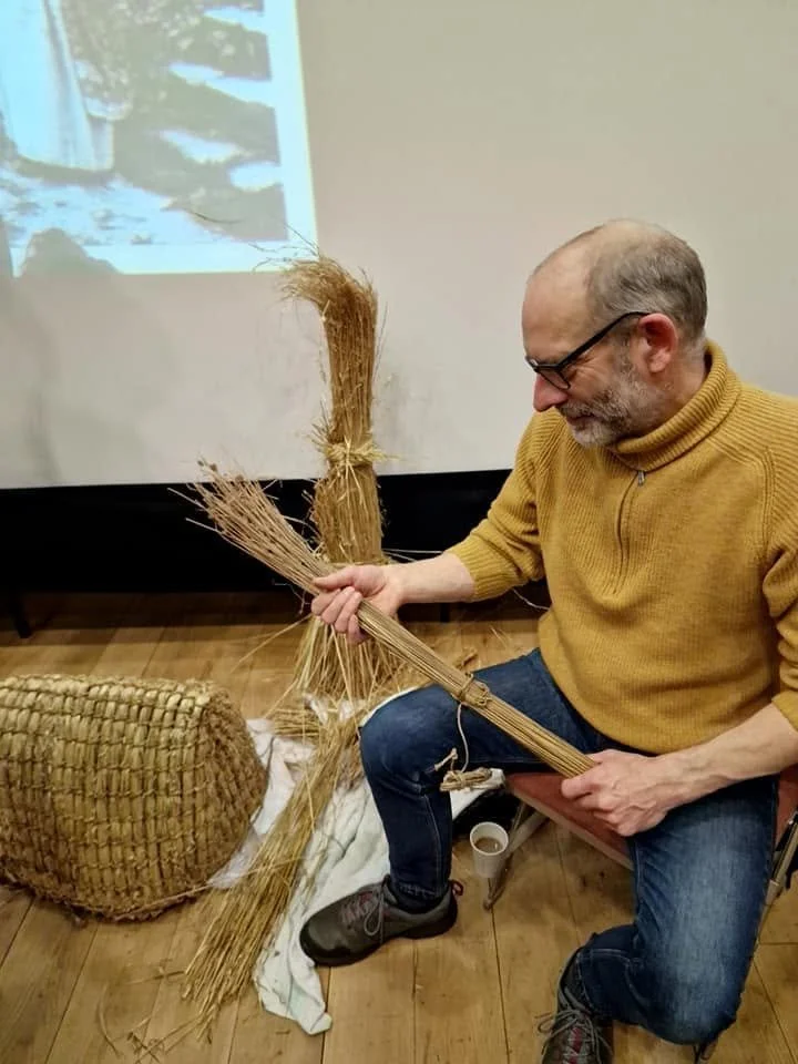 A man sitting in a chair wearing a mustard yellow sweater and jeans, holding dried straw in his hands, with a large woven basket and a white cloth on the wooden floor beside him. There is a projection of a snow-covered scene on the wall behind him.