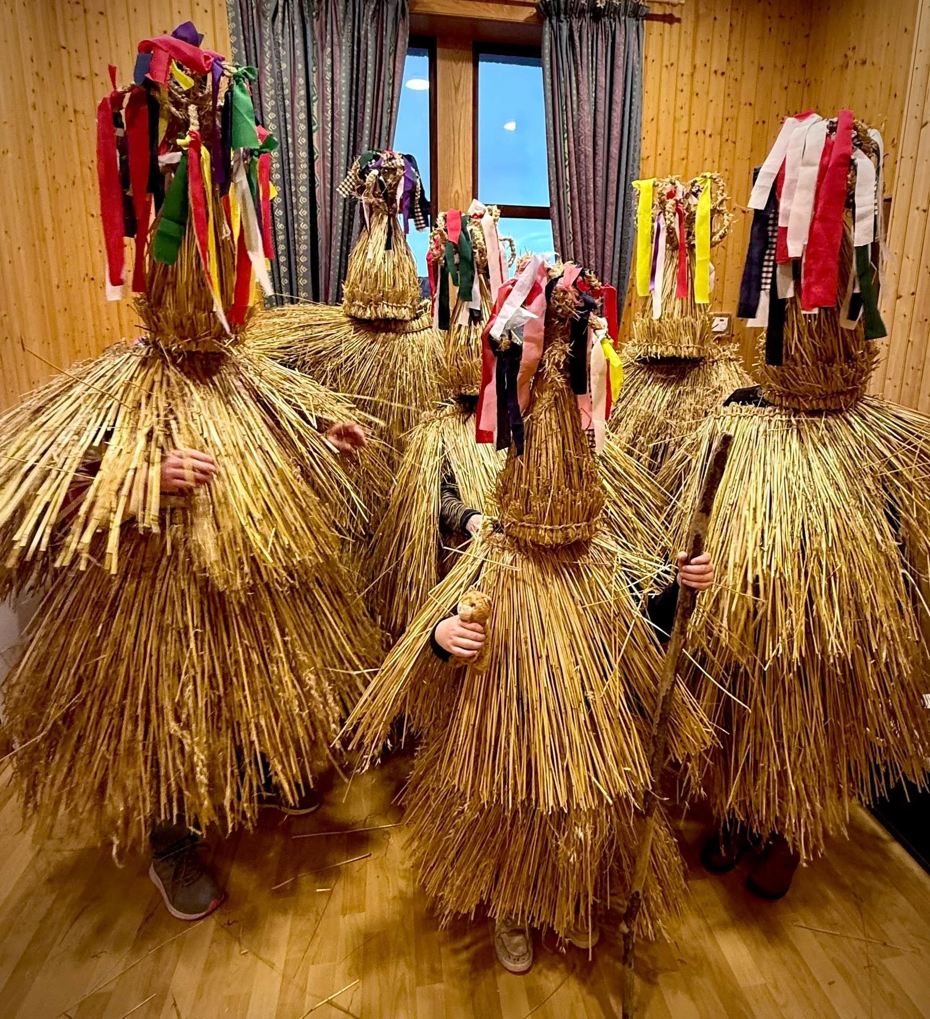 People dressed in straw costumes with colorful ribbons, standing inside a wood-paneled room with a window and floral curtains.