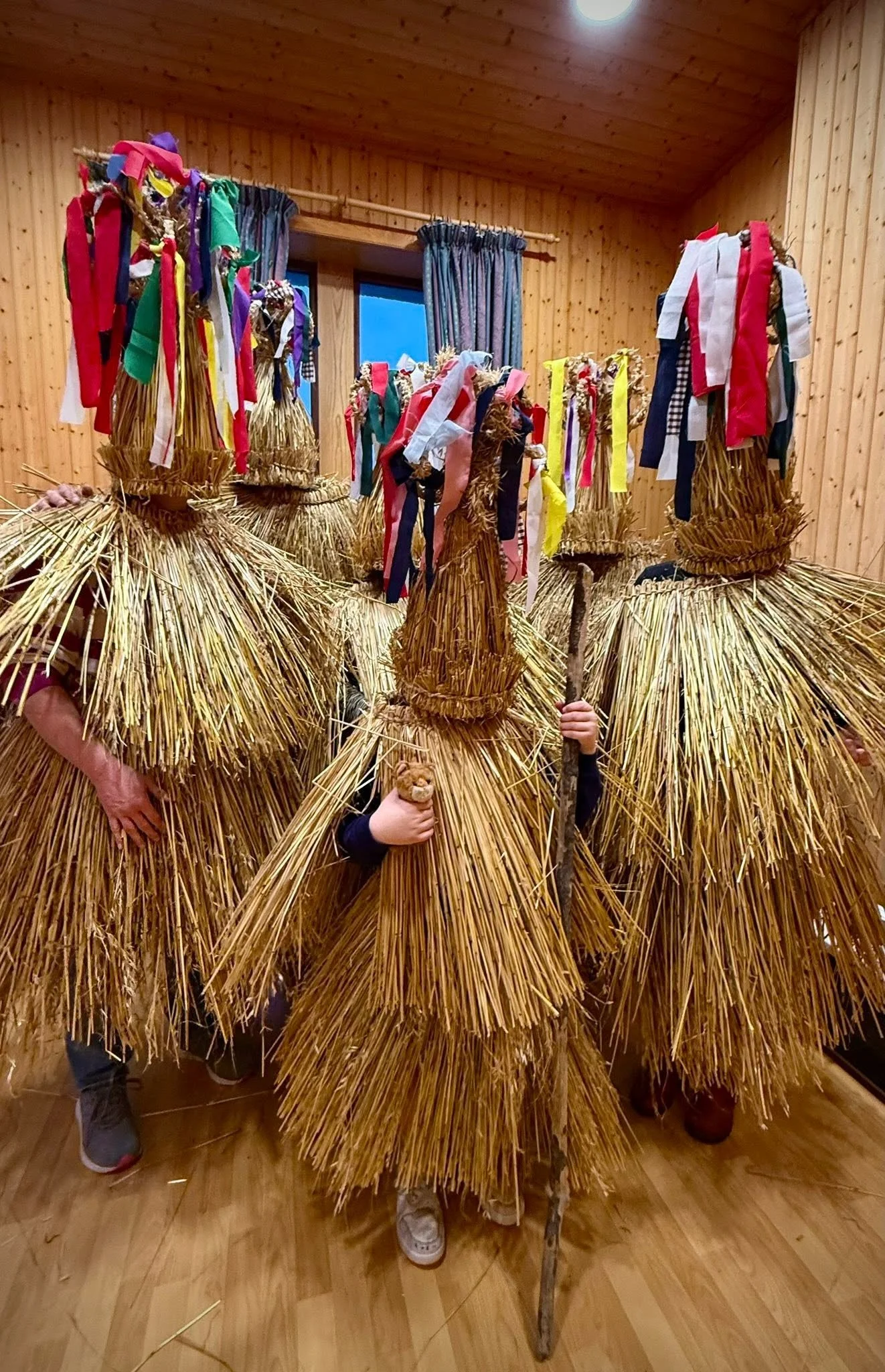 People dressed in traditional straw costumes with colorful ribbons and holding wooden sticks, standing on a wooden floor in front of a wooden-paneled wall, in a room with a window and curtains.