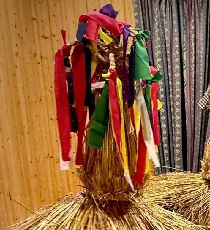 A decorative scarecrow made with a bundle of straw, dressed with a headpiece adorned with colorful fabric strips in red, yellow, green, purple, and white, set against a wooden wall and patterned curtains.