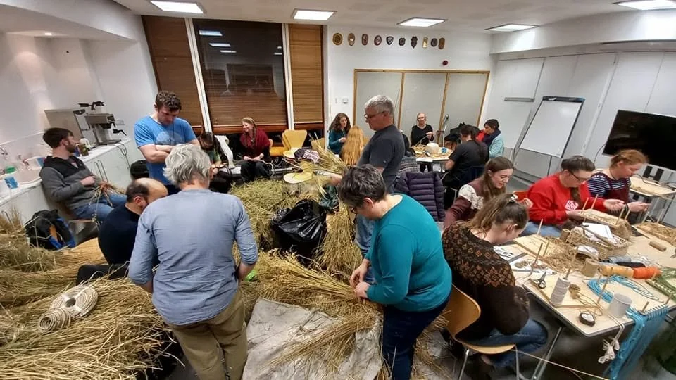 People working together in a classroom assembling straw and wicker items, with various tools and materials on the tables.