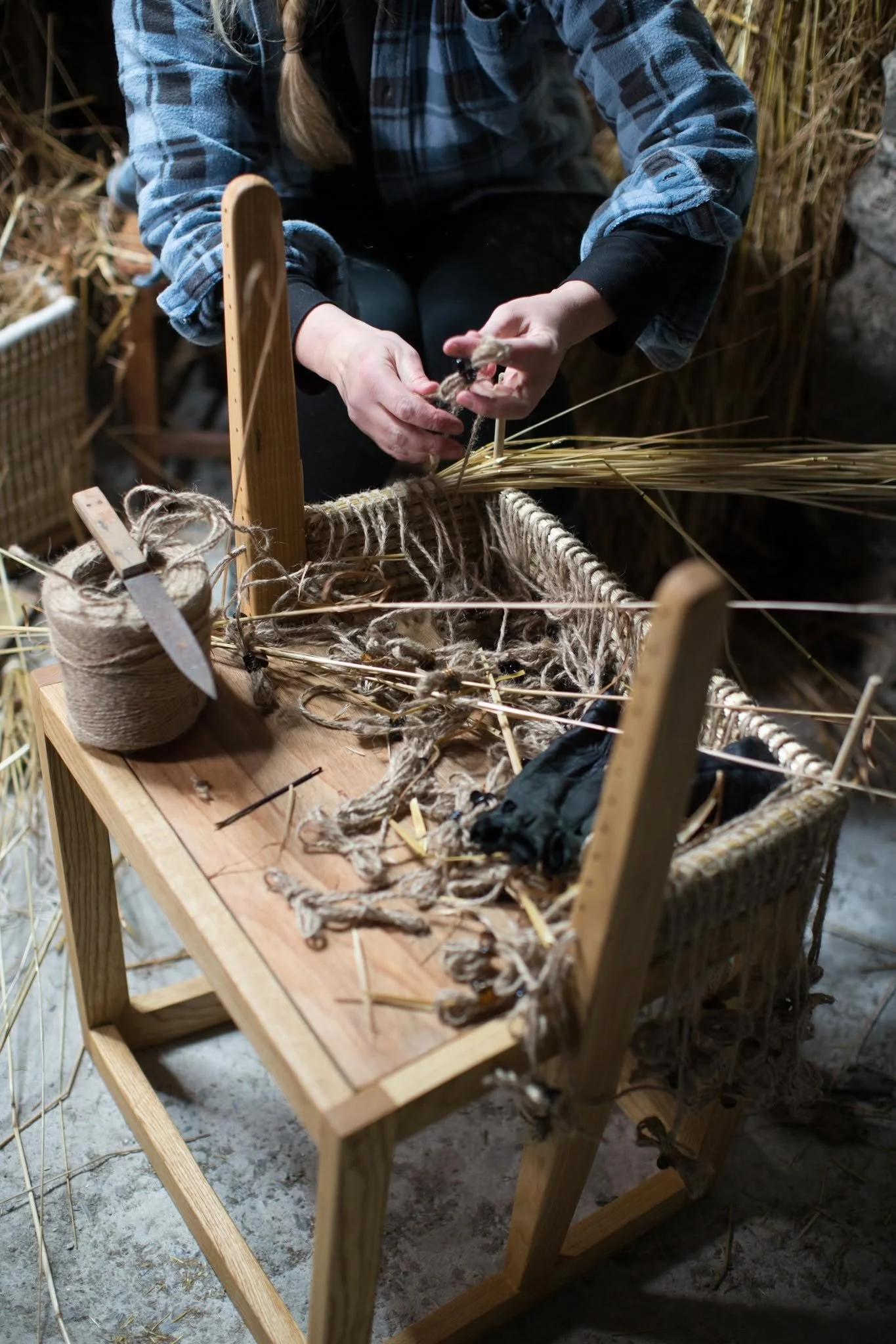 A person in a blue plaid shirt is weaving a basket using dried reeds or straw, with a partially completed basket in front of them on a wooden table.