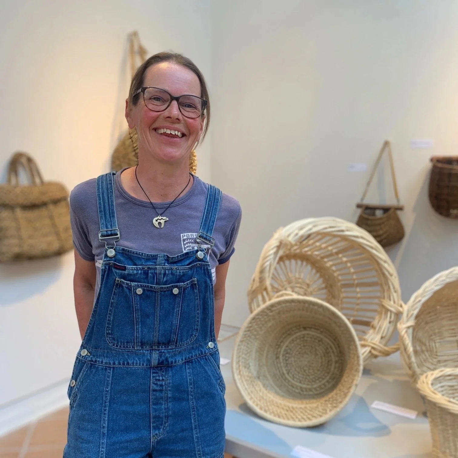 Woman smiling in front of a display of woven baskets at an art exhibit or craft fair.