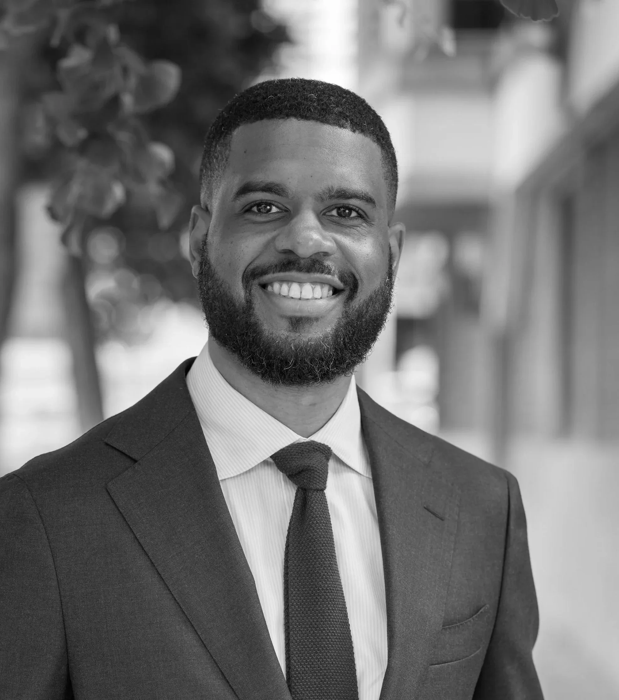 Close-up black-and-white photo of a smiling man with a beard, dressed in a suit and tie, outdoors with blurred trees in the background.
